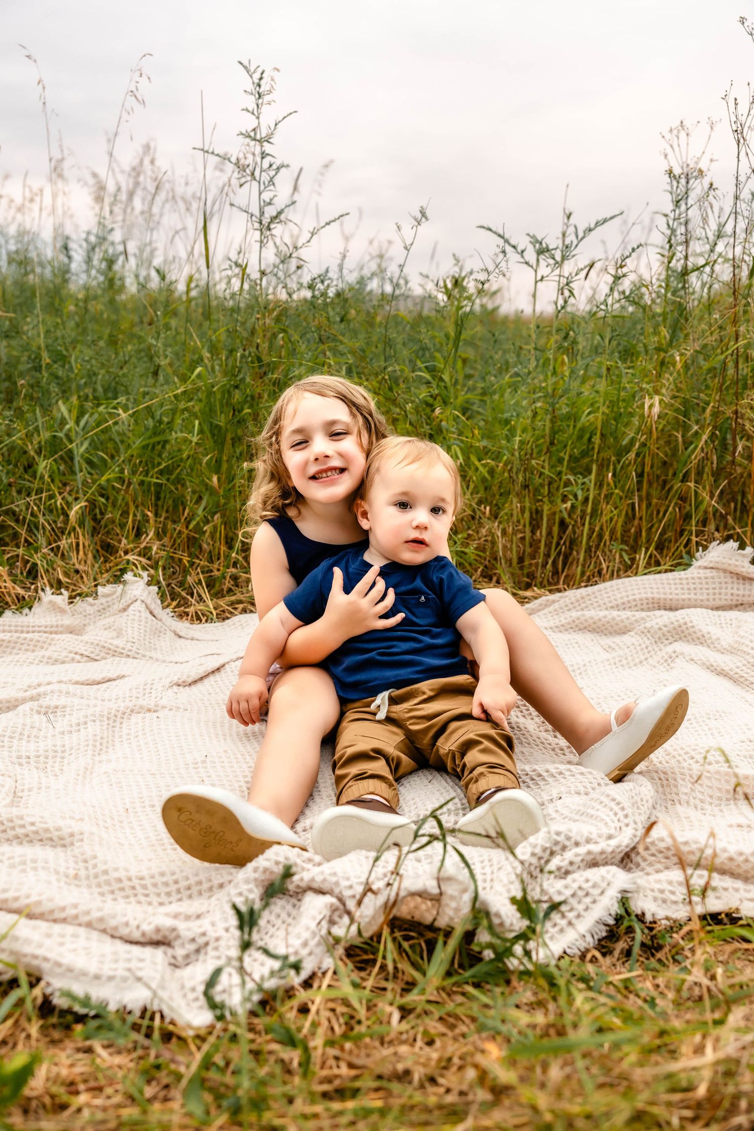 Two children sit together on a blanket in a field of tall grass on a cloudy day.