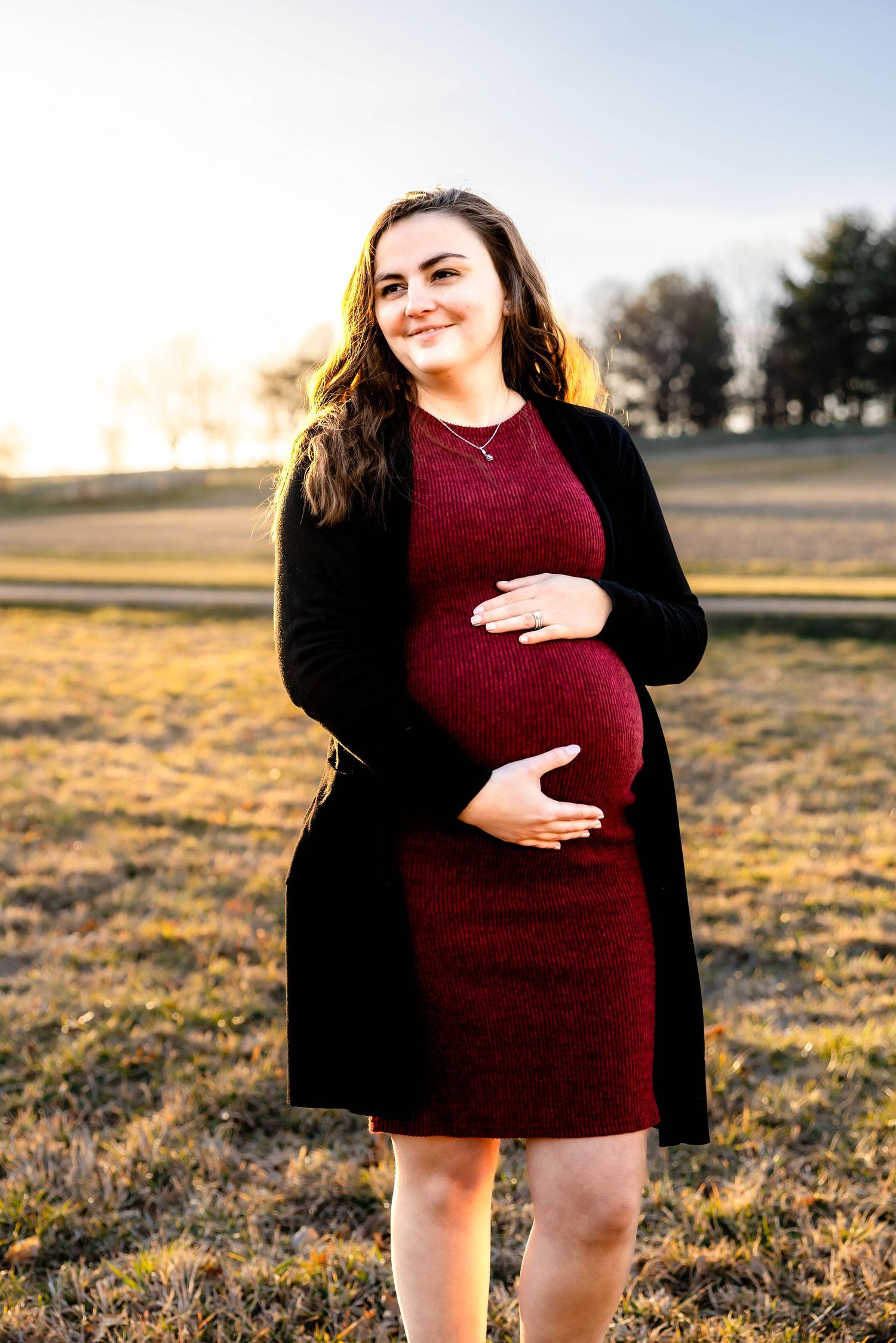 Pregnant woman in burgundy dress poses outdoors during golden hour.