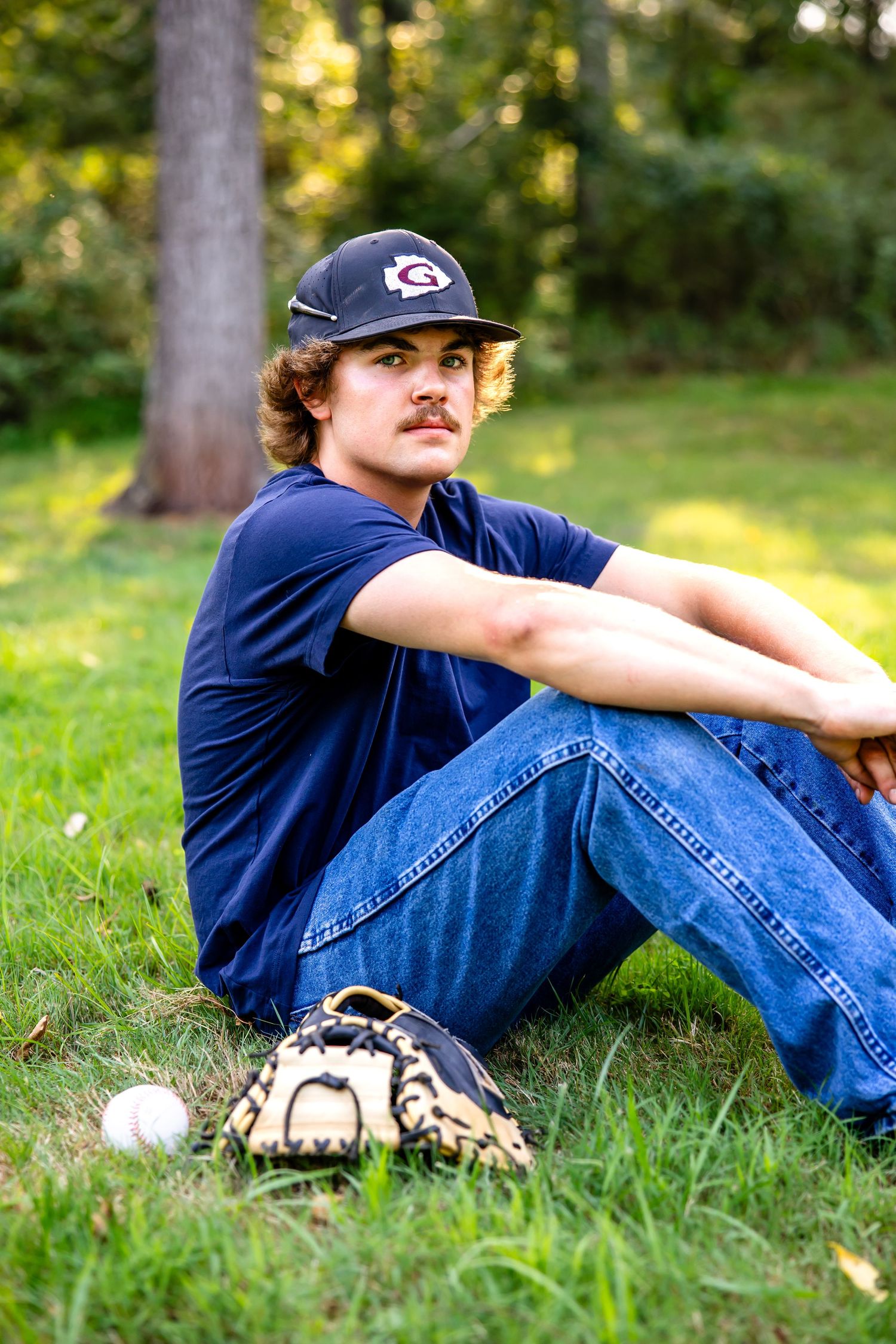 Baseball player in casual attire sitting on grass with baseball gear nearby.