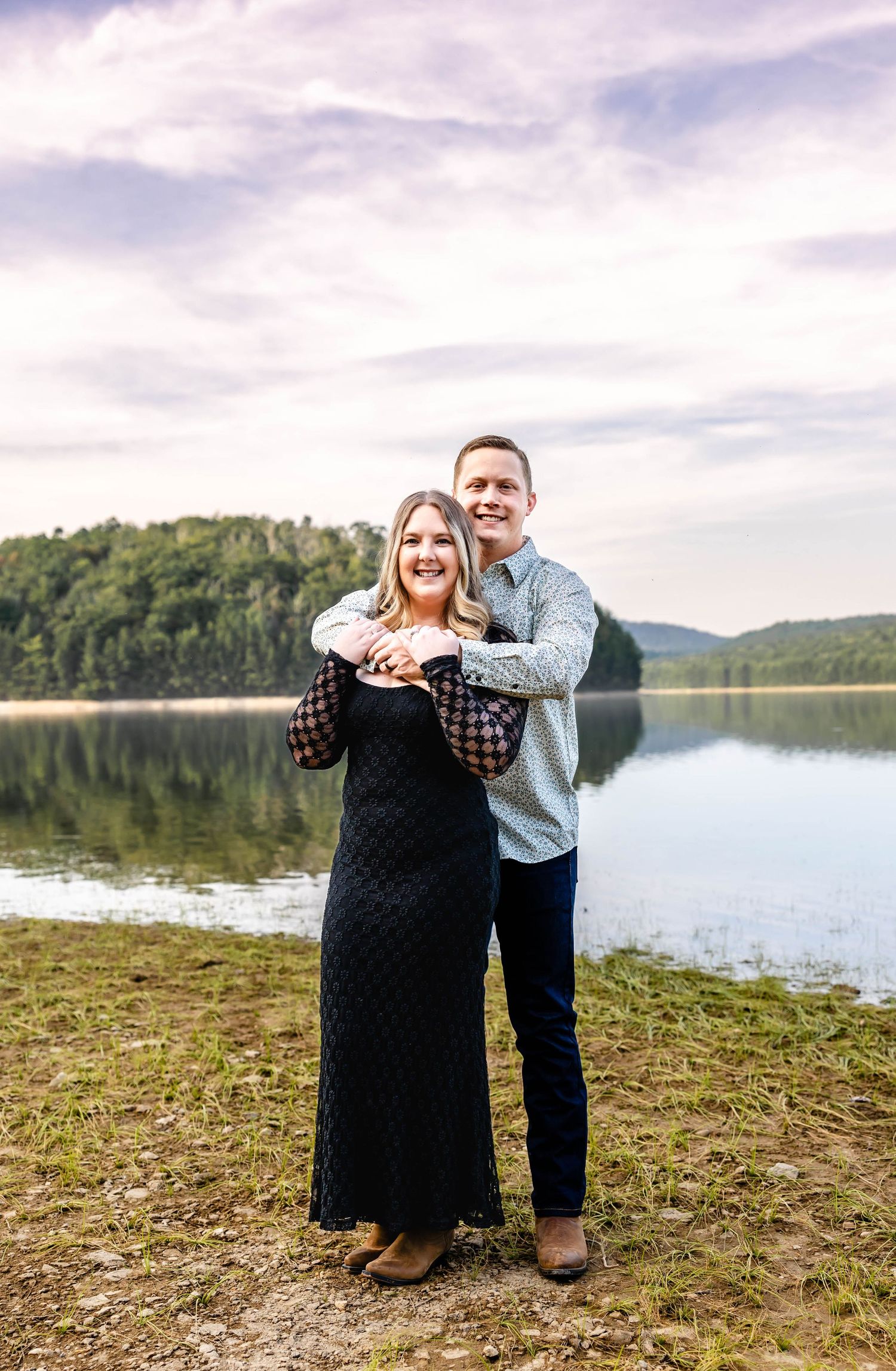 A couple embraces by a peaceful lake with rolling hills and clouds in the background.