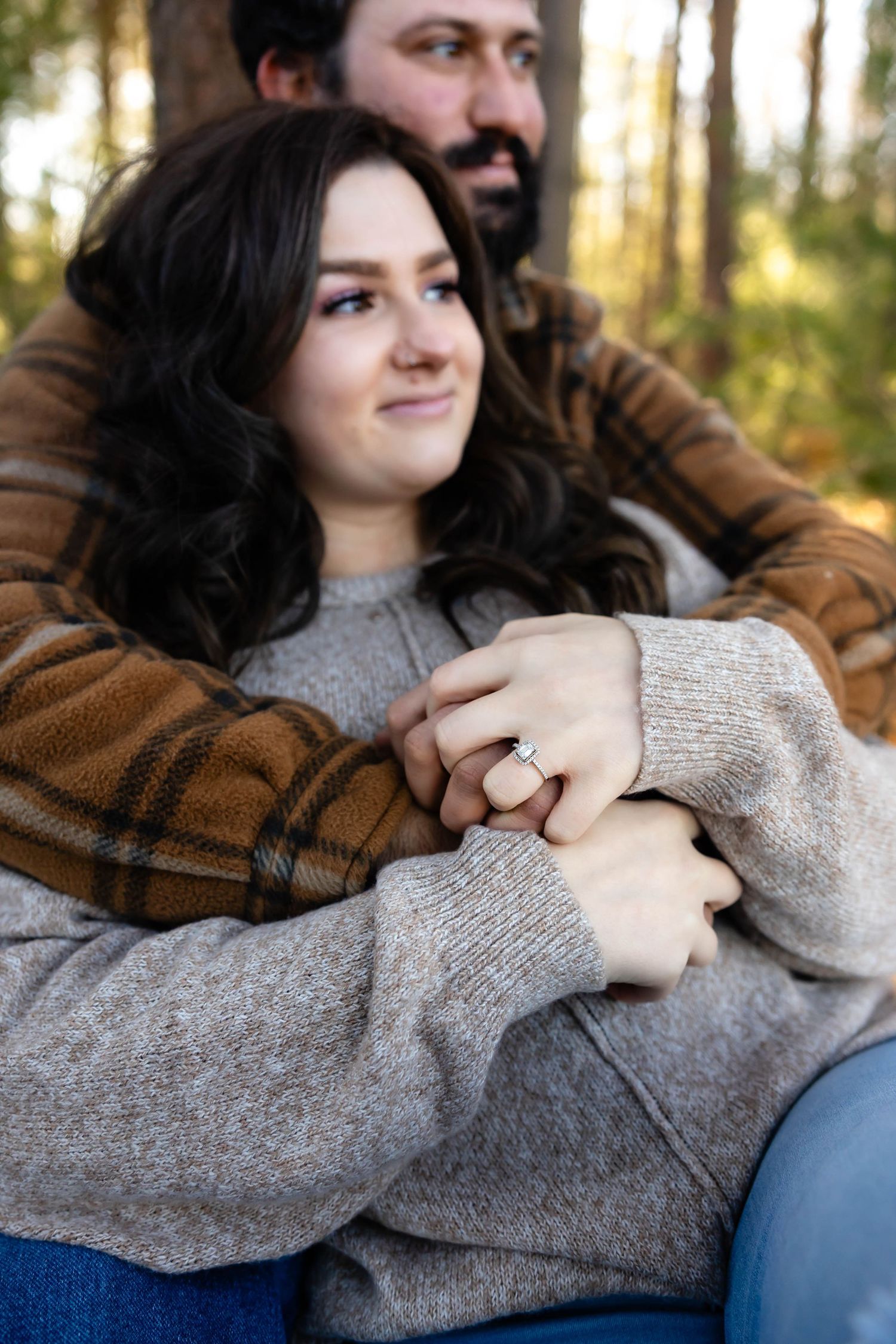 A couple snuggles close while sitting in a forest, showing off an engagement ring.
