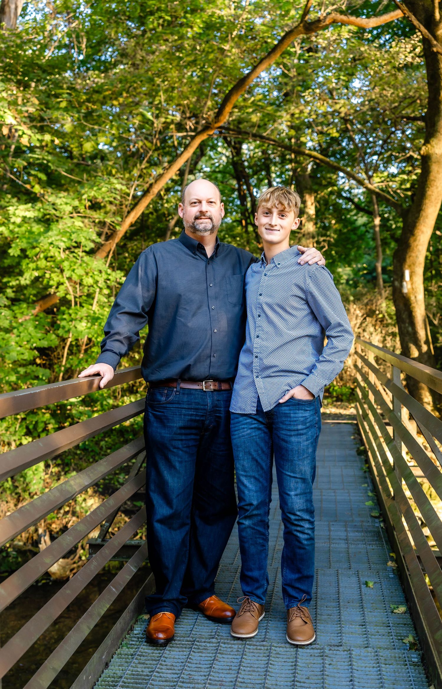 Two people in blue casual wear stand together on wooden bridge path surrounded by trees.