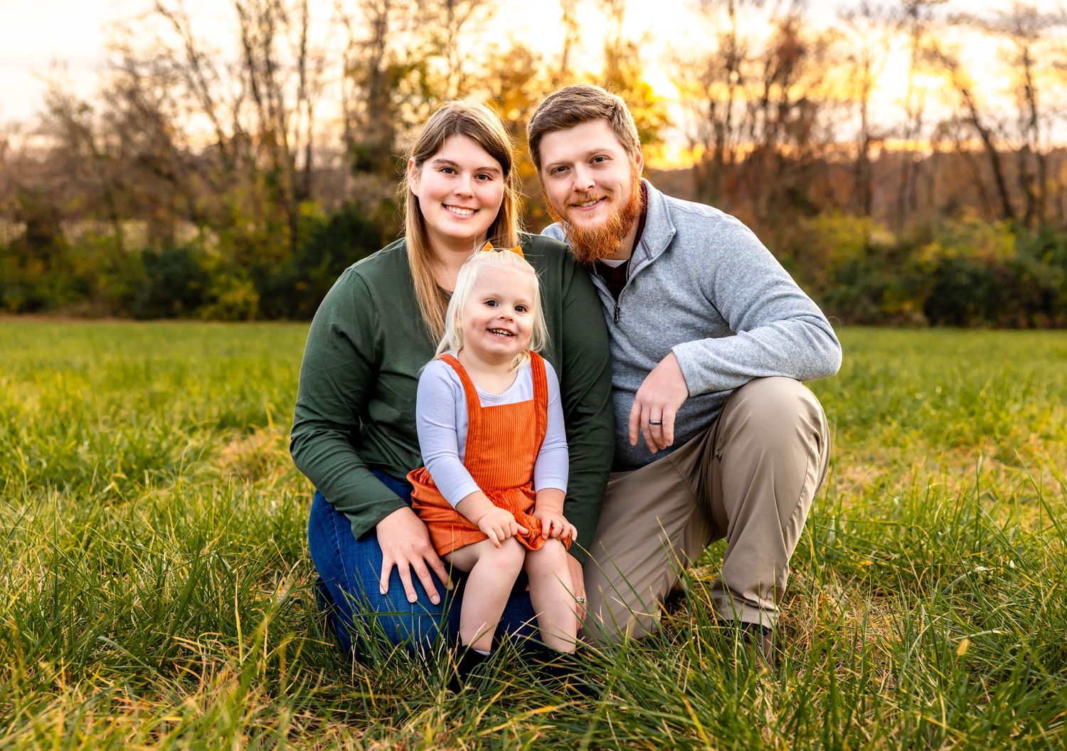 A family portrait taken in a grassy field during golden hour with vibrant fall colors in the background.