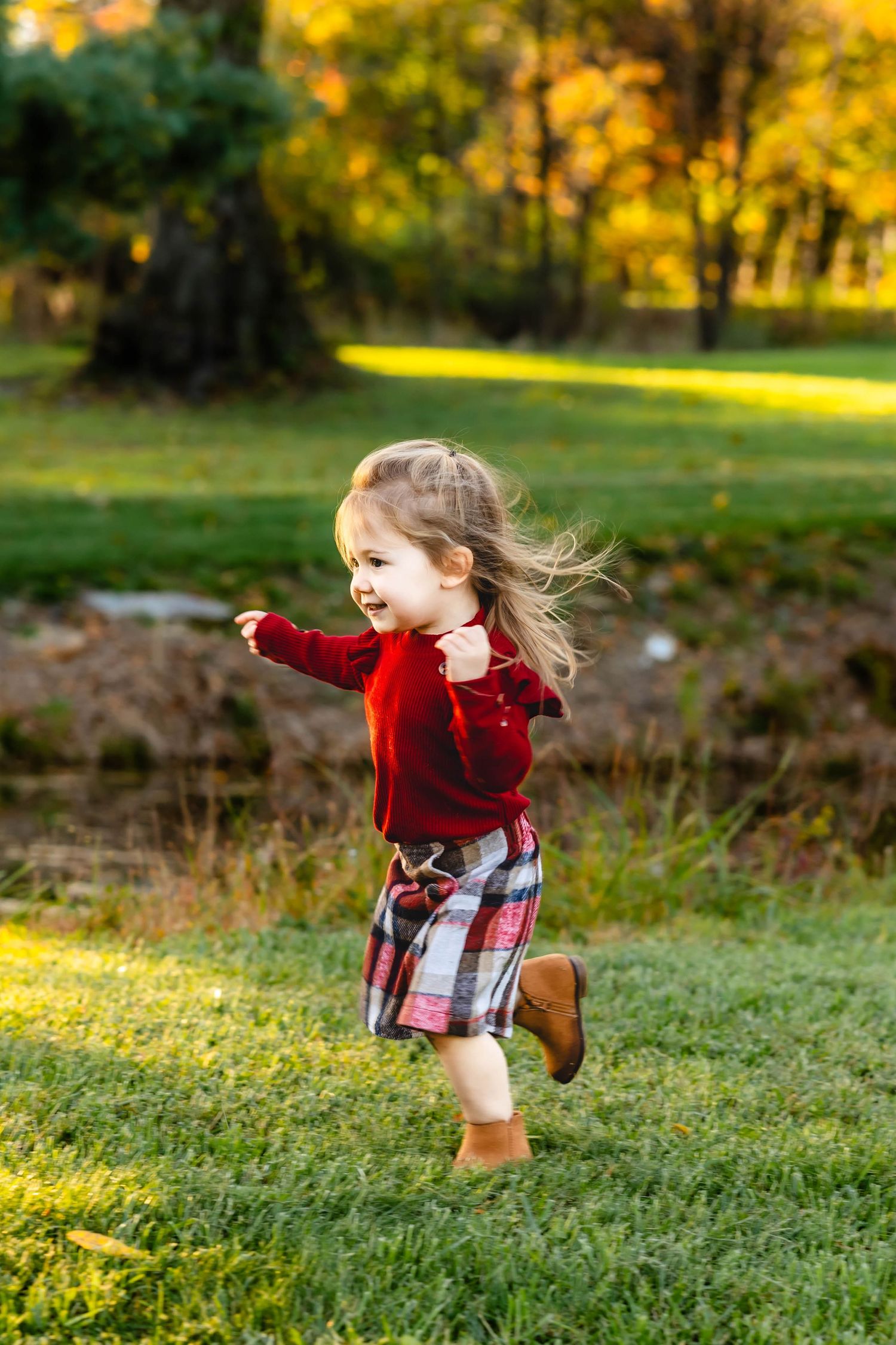 Child in red velvet top and plaid skirt runs through grass during golden autumn afternoon.
