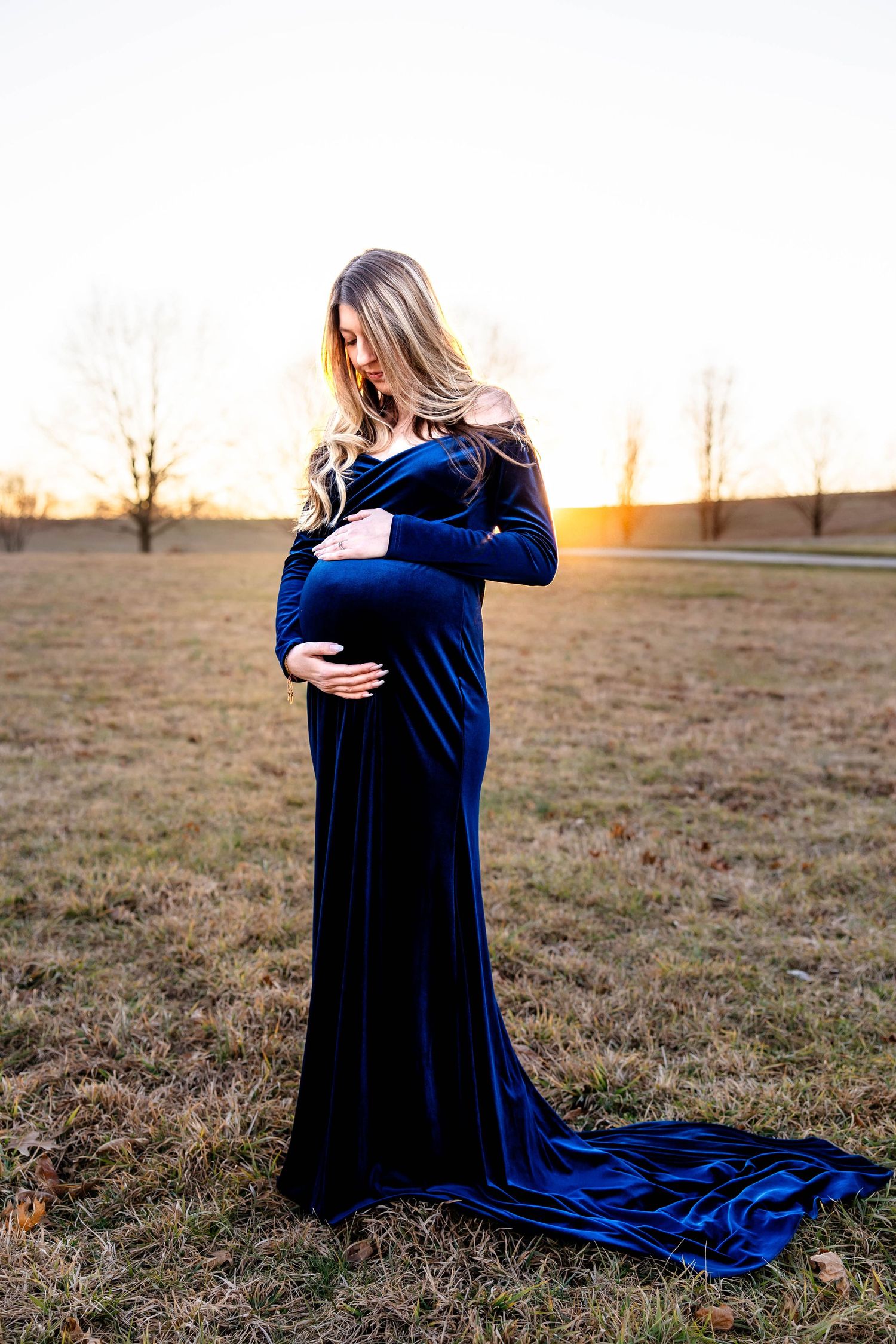 Expecting mother in flowing navy velvet gown poses in field at sunset.