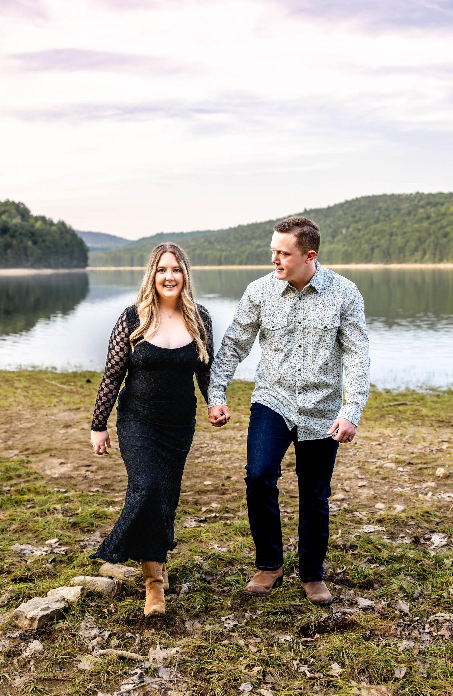 A couple walking hand in hand along a lake shore with mountains in the background.
