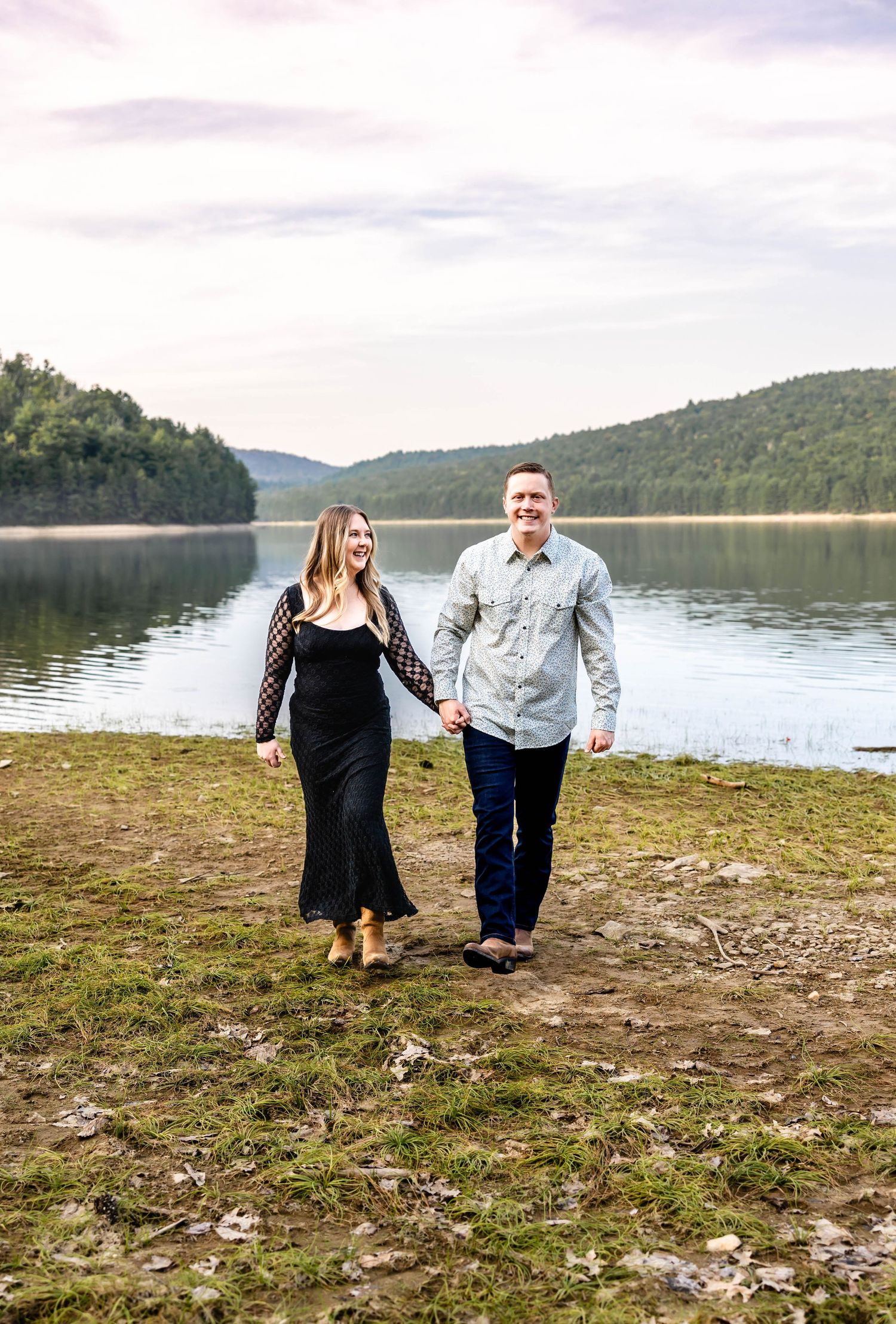 Couple walks hand in hand along shoreline with mountain lake backdrop.