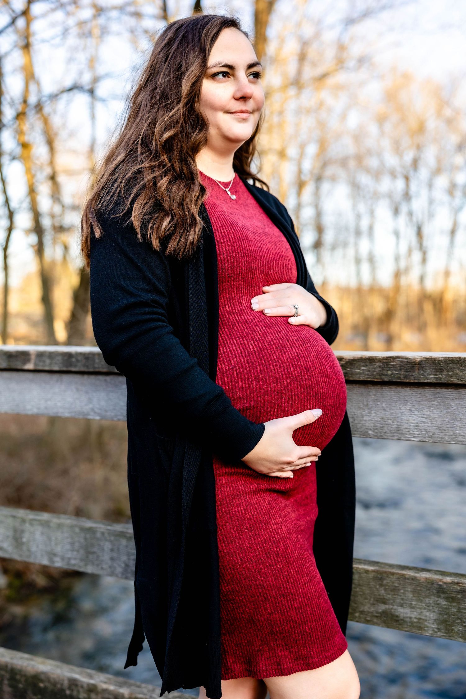 An expectant mother in a burgundy dress poses against an autumn landscape with a stream.