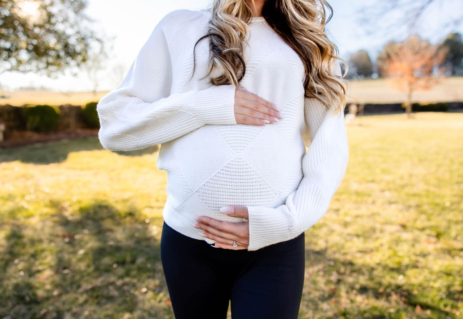 Close up view of a pregnant belly in white knit sweater against golden autumn backdrop.