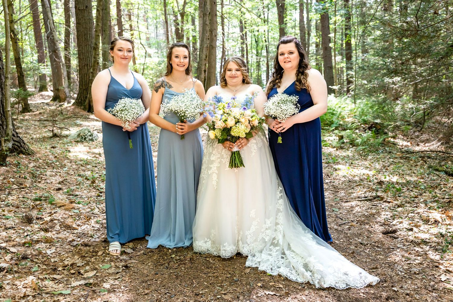 Wedding party in blue and gray dresses poses in wooded setting with bride in lace gown.