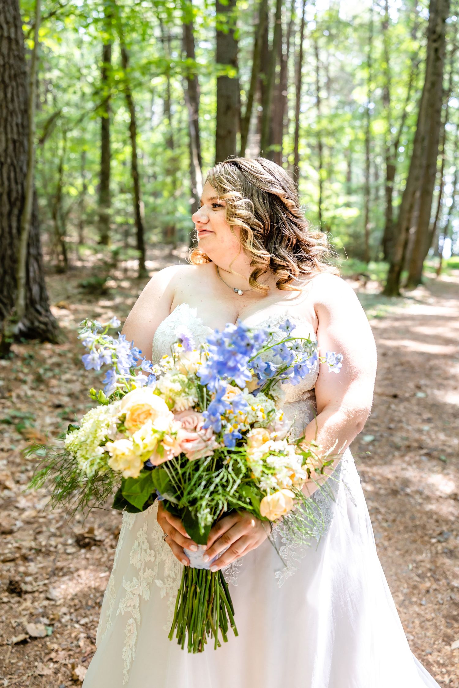A radiant bride with a stunning floral bouquet poses in a sunlit forest setting.