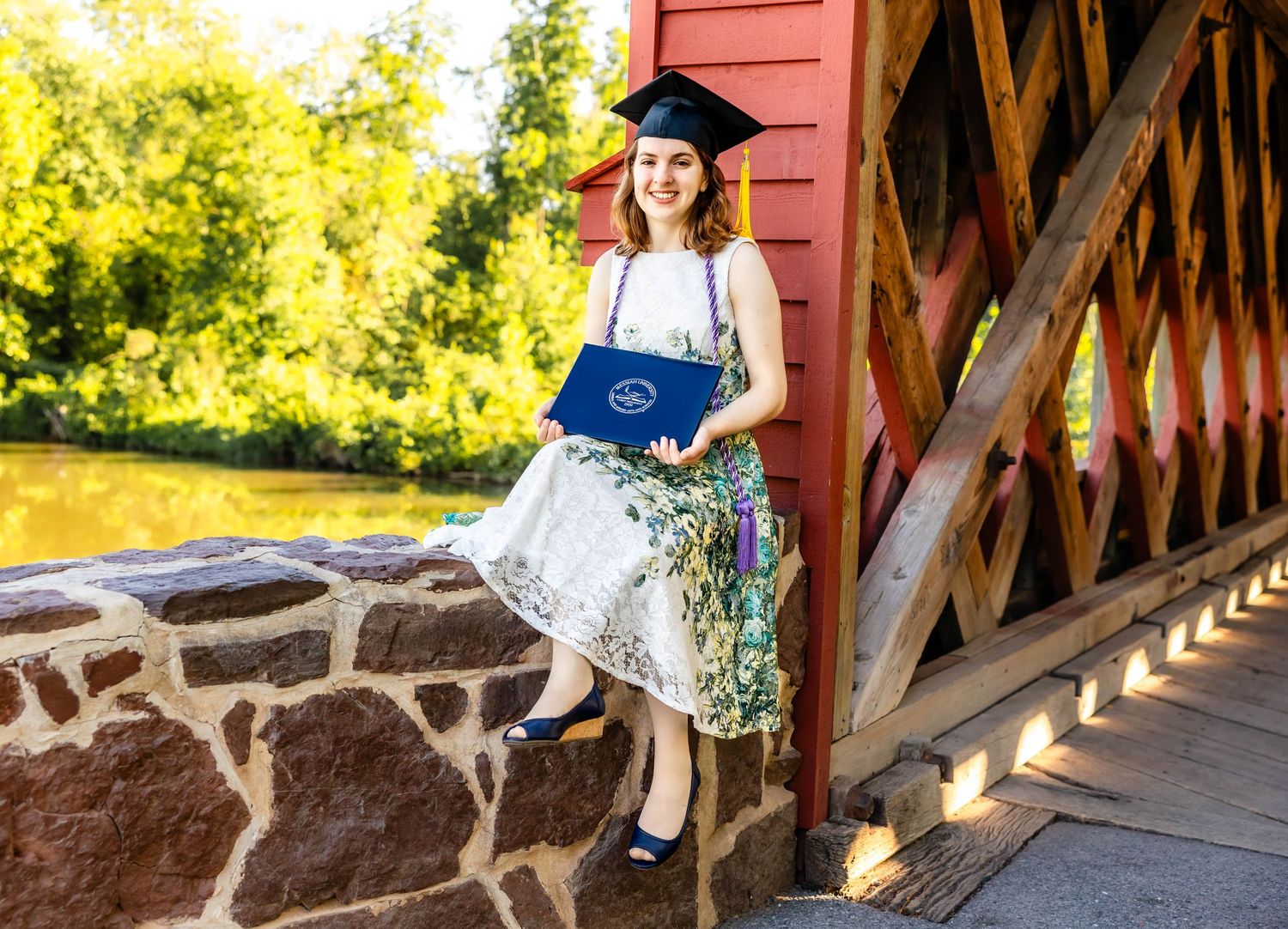 A graduate in cap and gown poses on a covered bridge with rustic stone foundation.