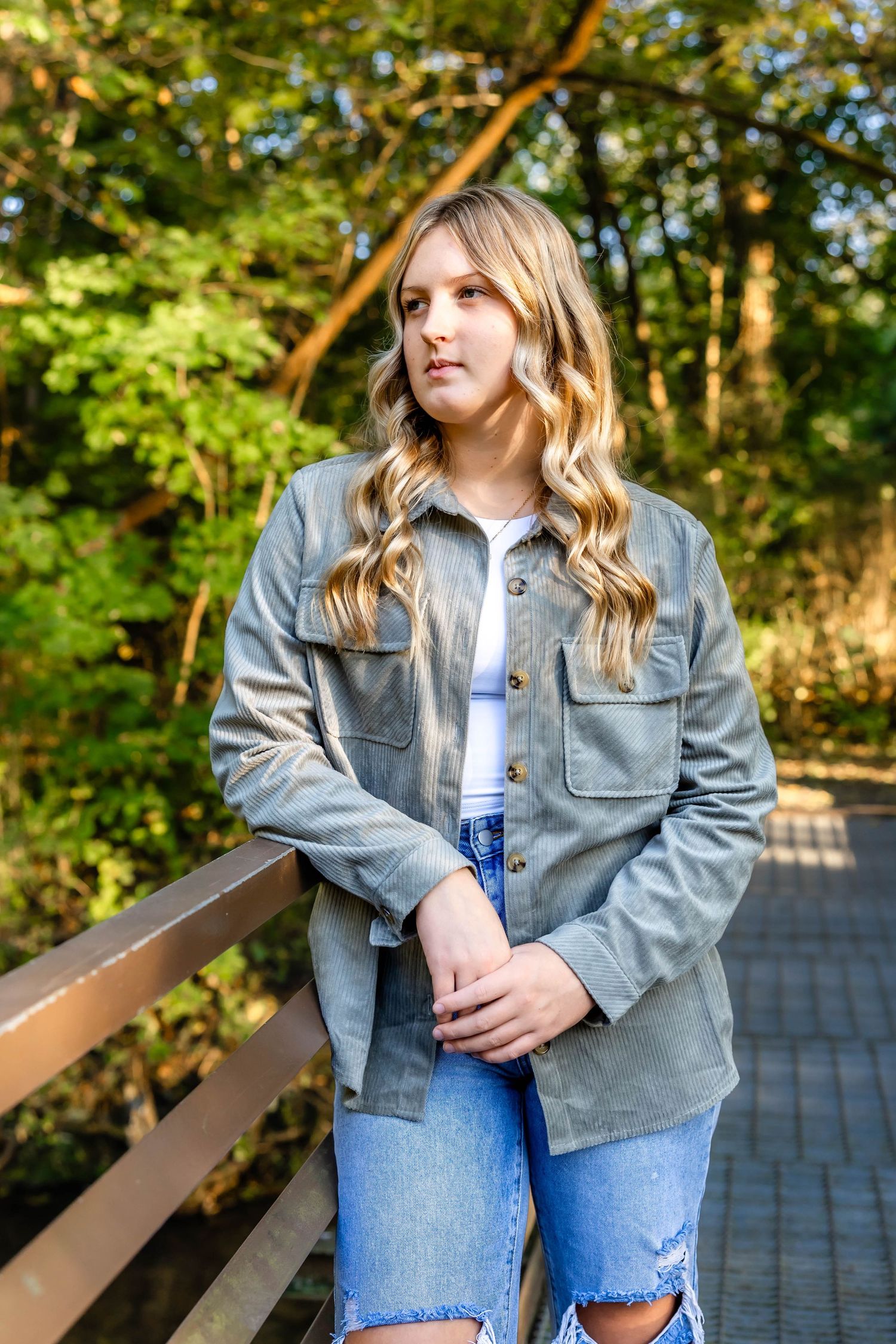 Person wearing gray denim jacket and ripped jeans stands on wooden boardwalk surrounded by lush greenery.