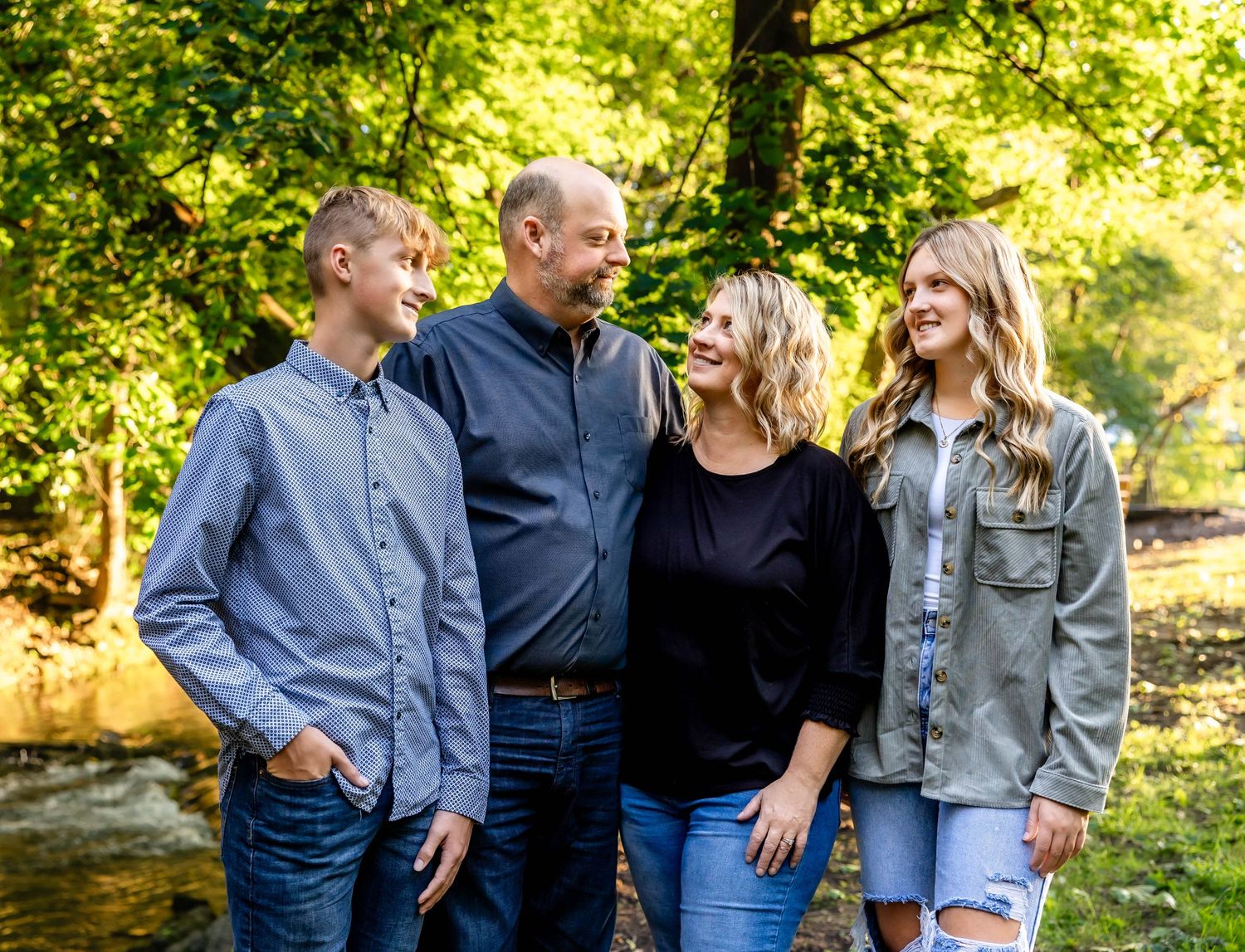 A family of four stands together outdoors with sun-dappled trees in the background.