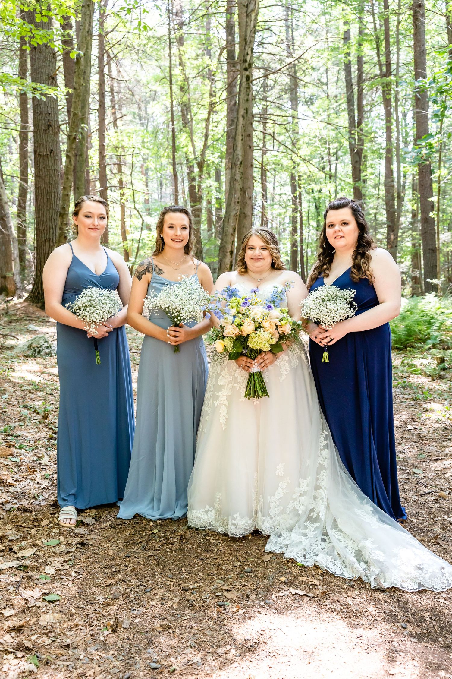 Four bridesmaids in varying shades of blue holding bouquets pose in a forest setting.