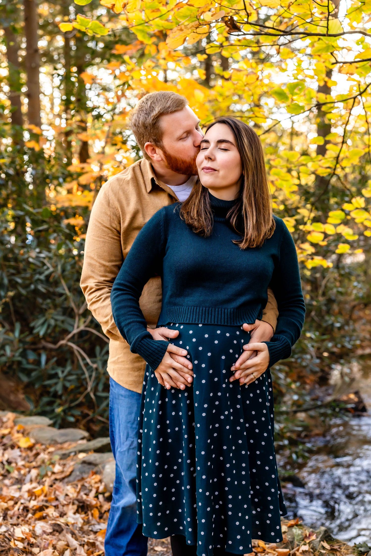 Couple sharing tender moment among fall foliage with golden sunlight filtering through trees.