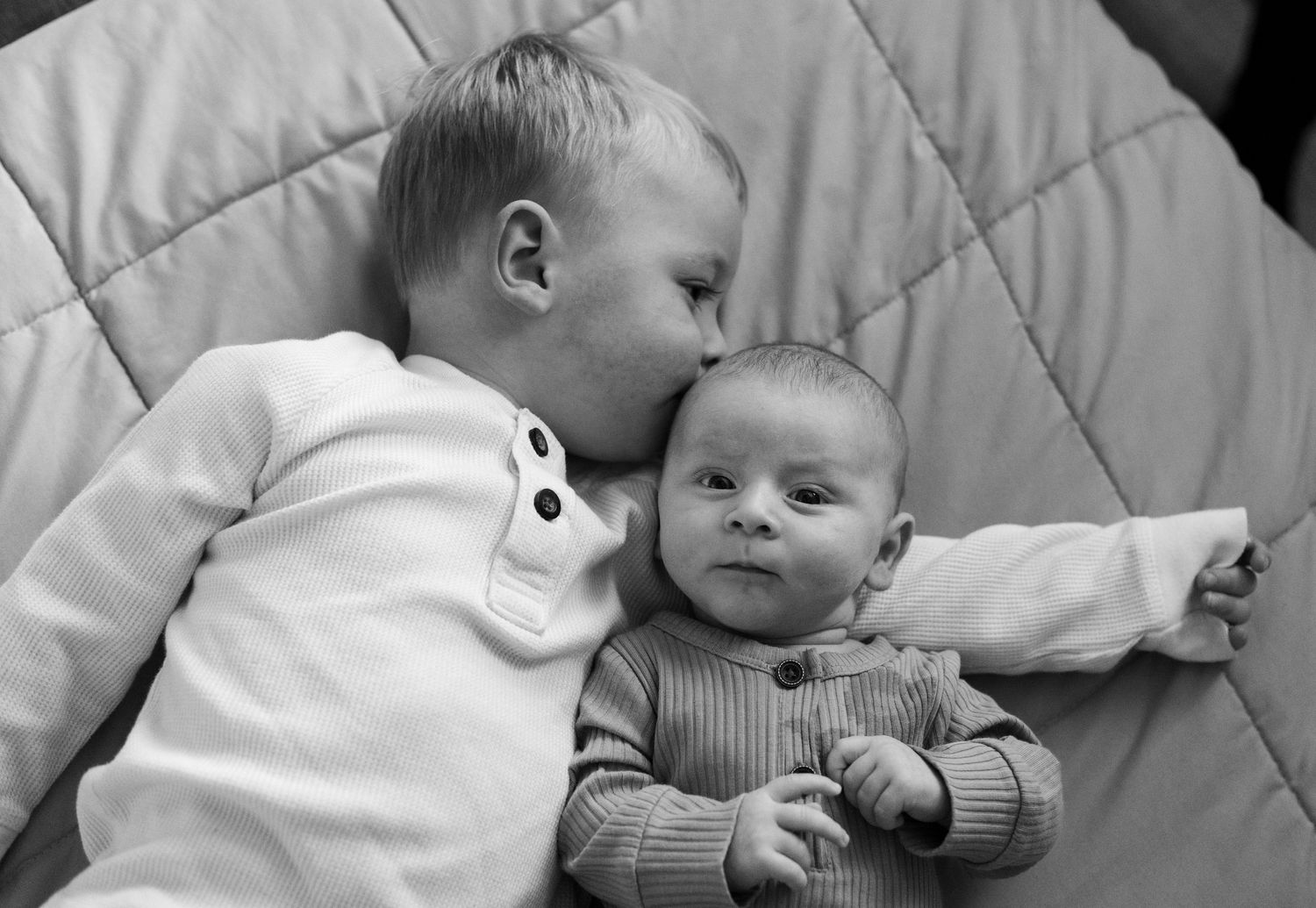 Black and white portrait of two siblings sharing a tender moment on a quilted blanket.