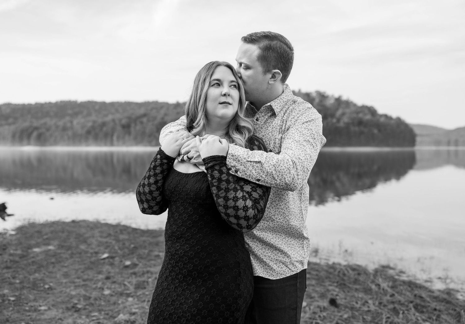Black and white portrait of couple embracing by serene lake with tree-lined shore.