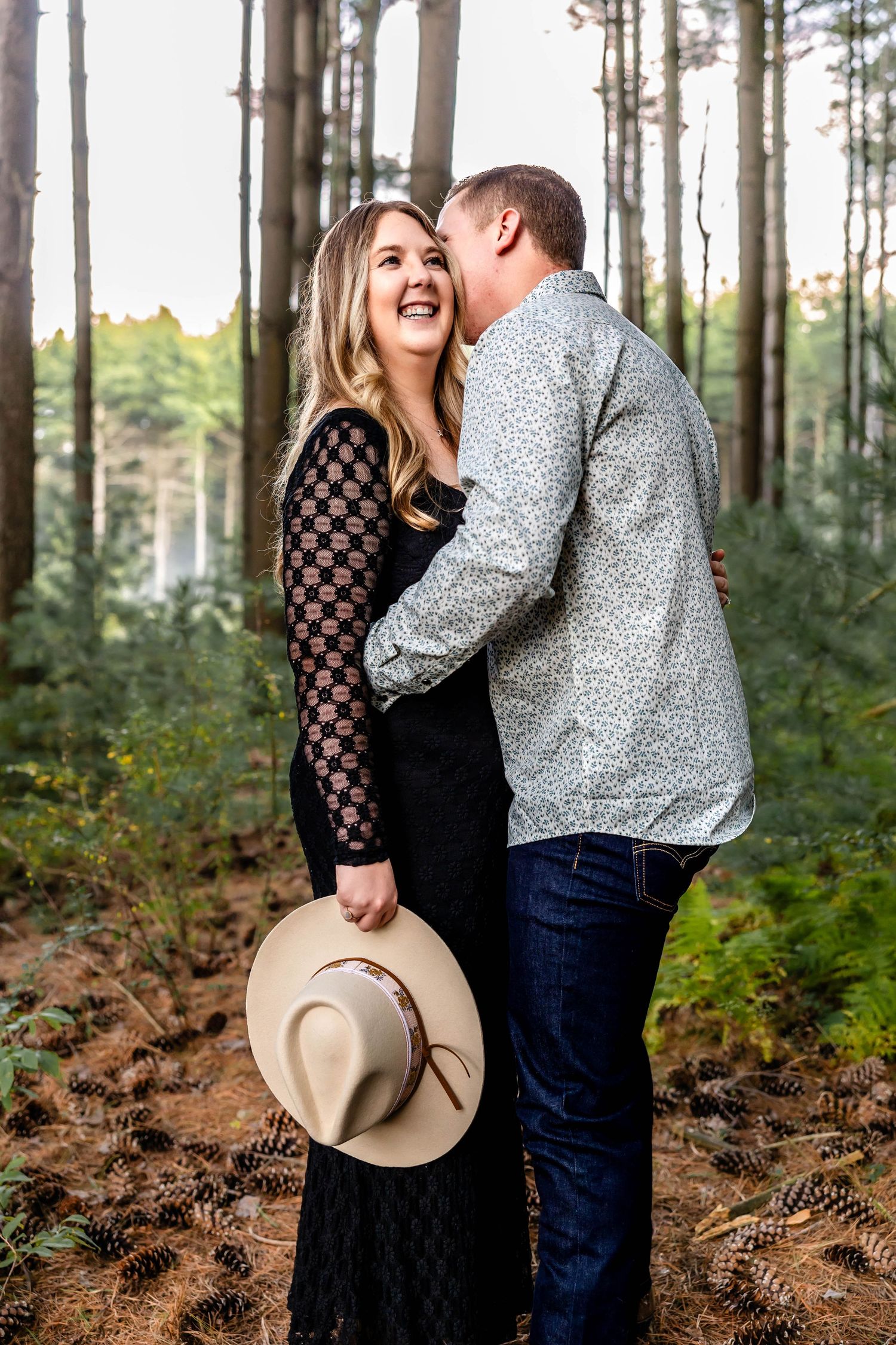 A playful couple wearing western hats shares a laugh in a pine forest.