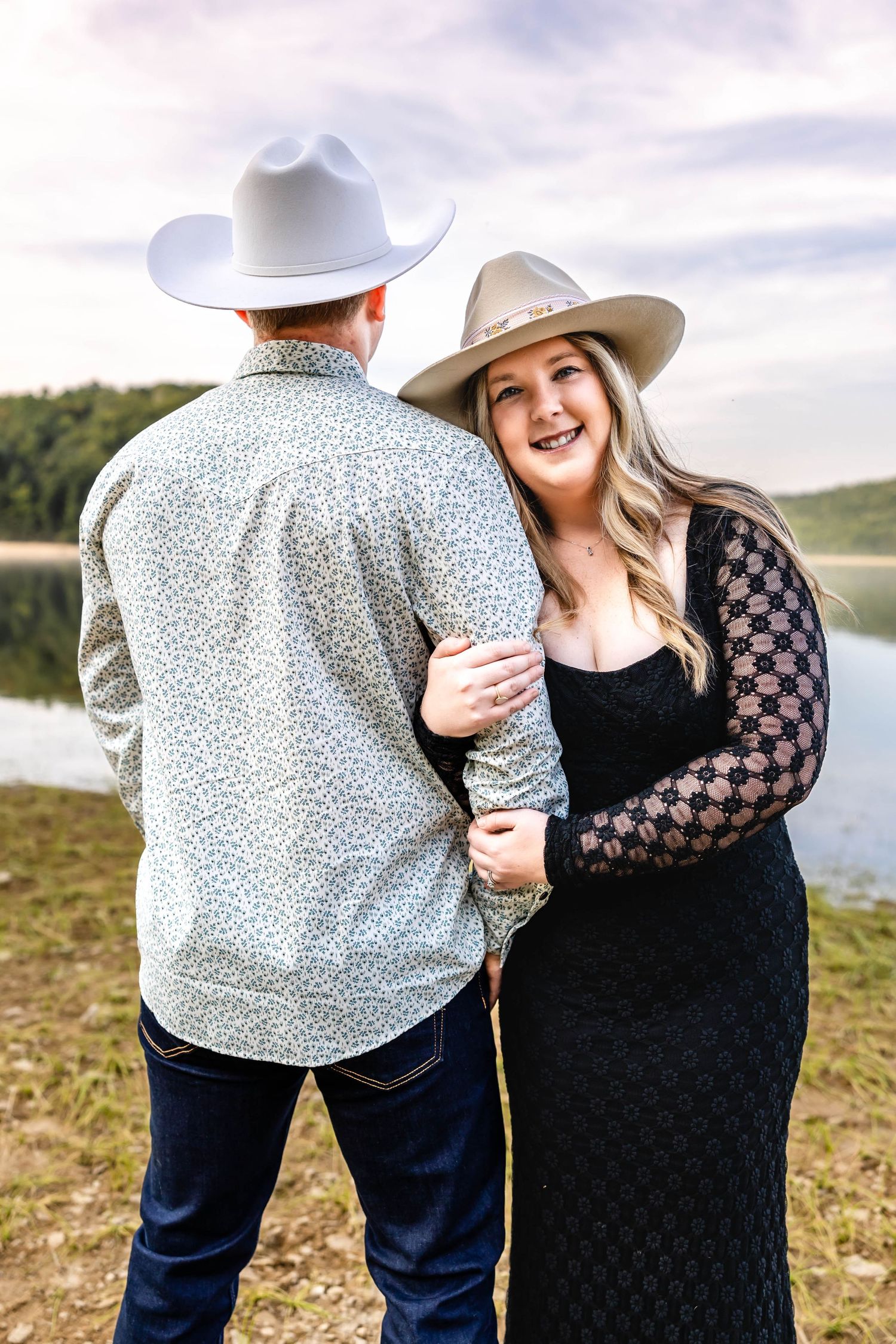 Couple in western attire shares tender moment by scenic lake during engagement shoot.