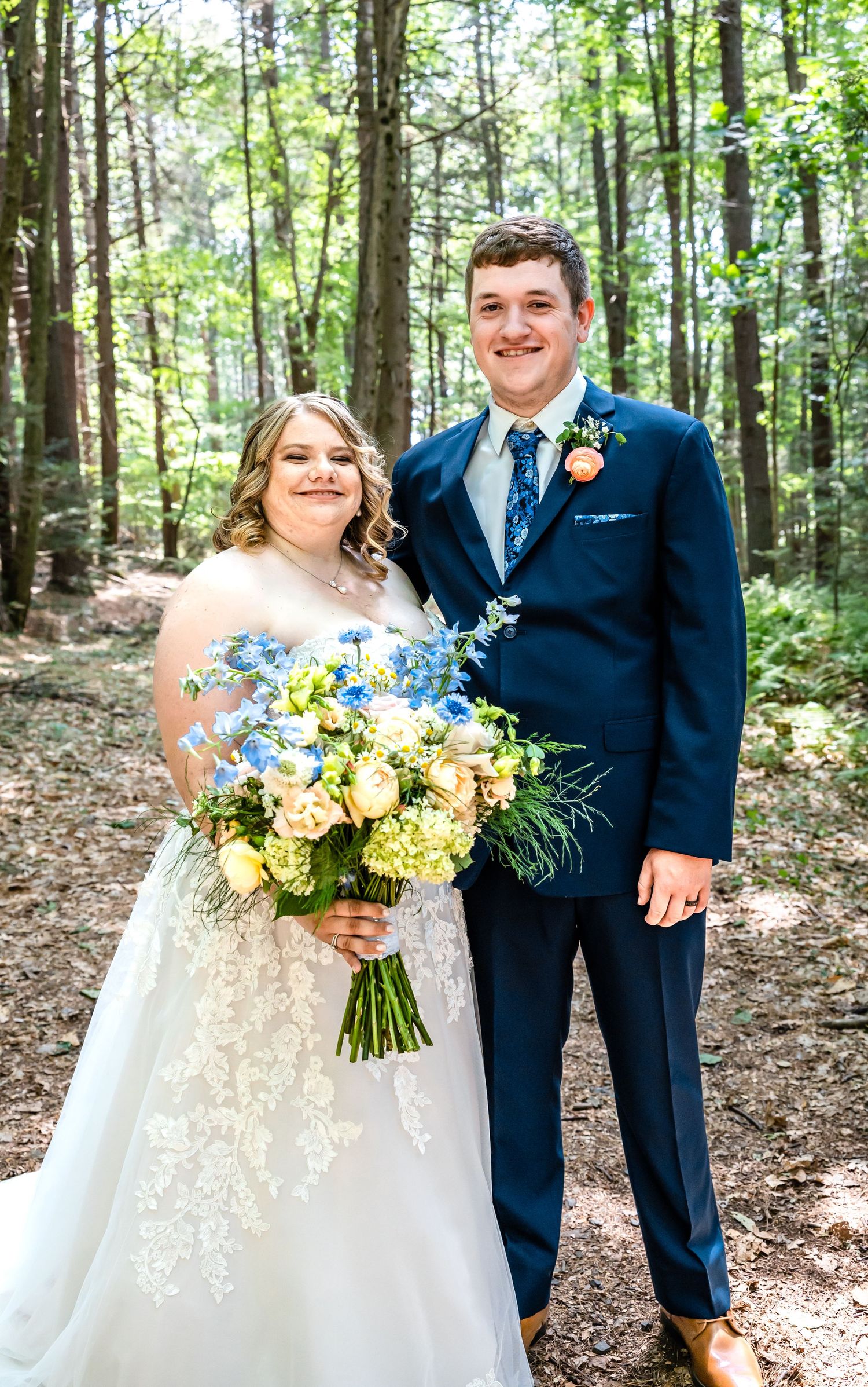 Newlyweds share joyful moment in forest setting with blue and yellow bouquet.
