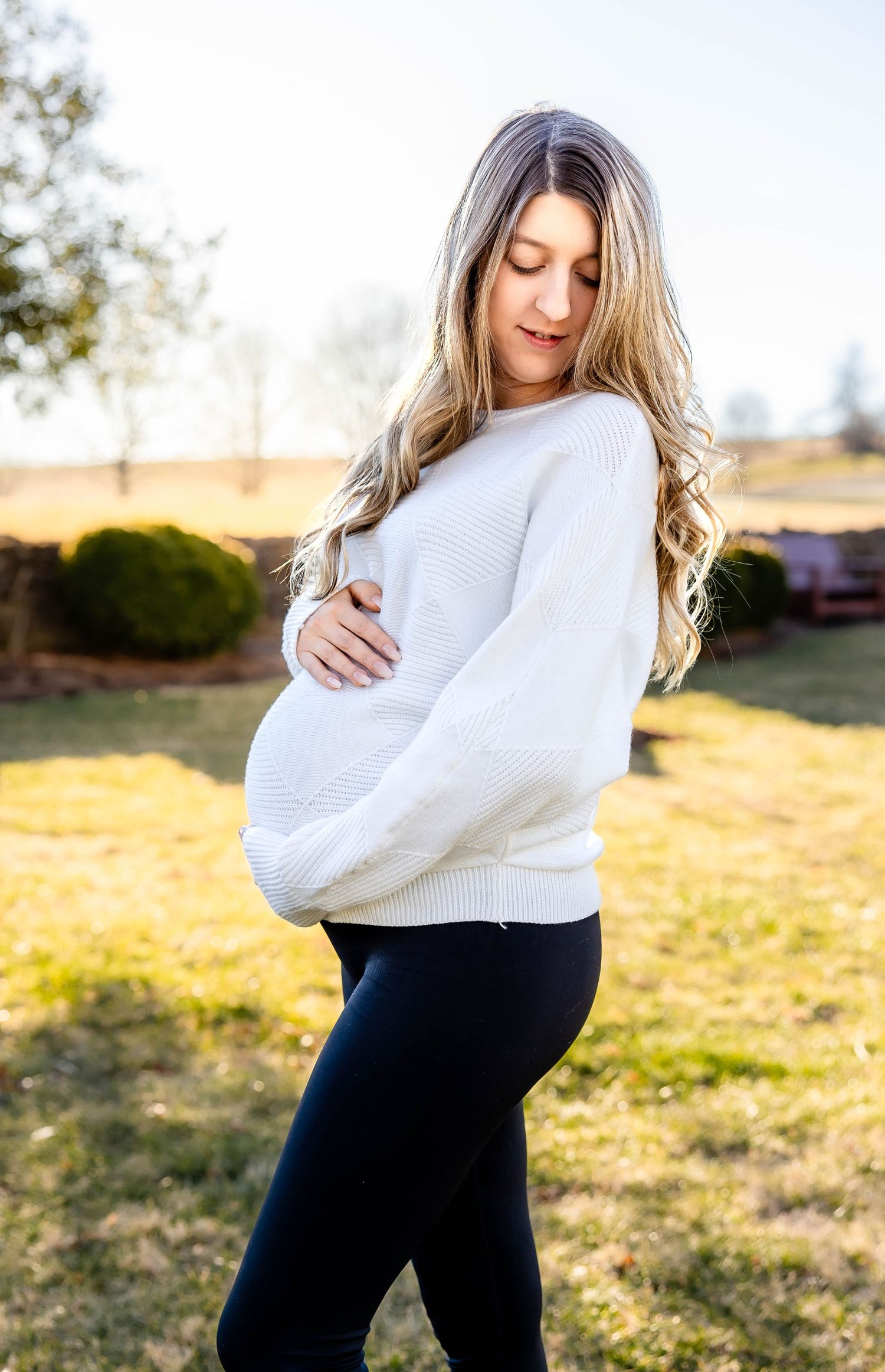 Maternity portrait of woman in white sweater holding her baby bump against sunlit outdoor background.