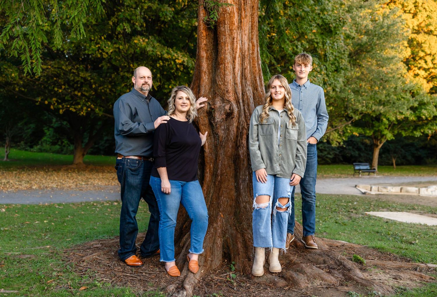 Family poses around large tree trunk in autumn park setting with fallen leaves.