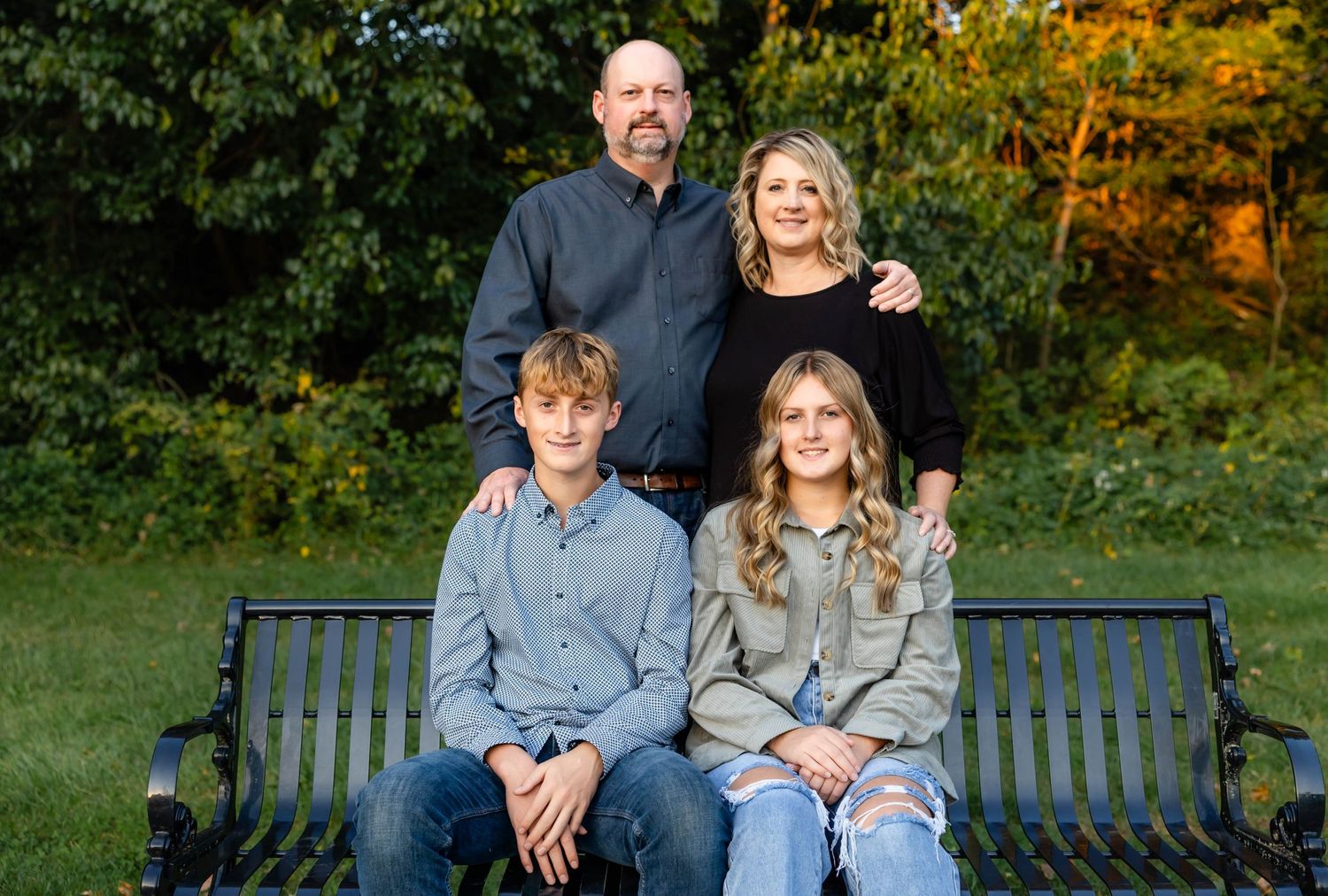 A family group sits together on a green metal park bench surrounded by autumn foliage.