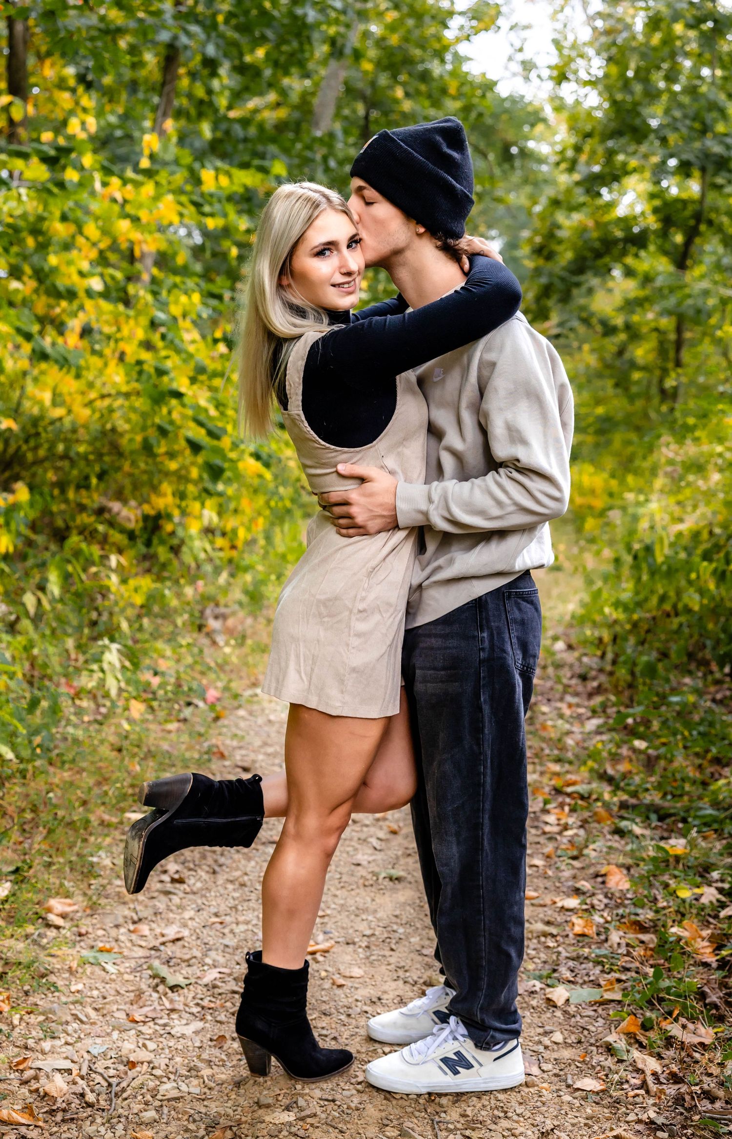 Stylish couple embracing on forest path with golden autumn leaves in background.