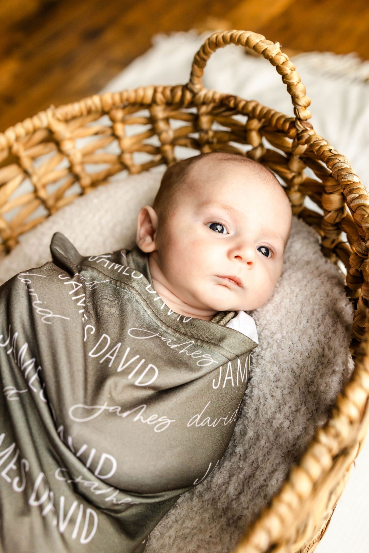 Close up of an infant resting in a woven basket wrapped in a personalized gray swaddle.