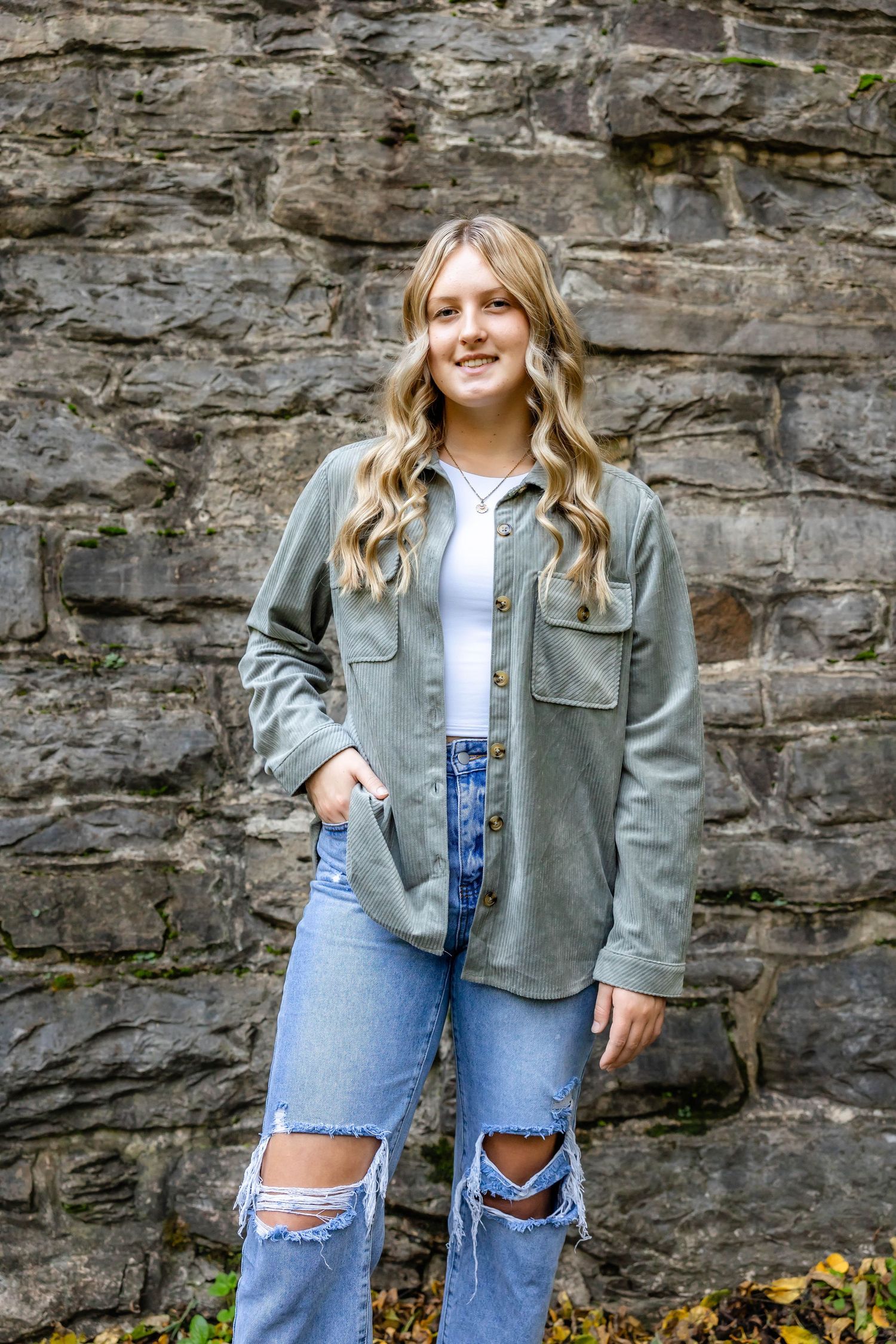 A teen in a gray jacket and distressed jeans poses against a stone wall outdoors.