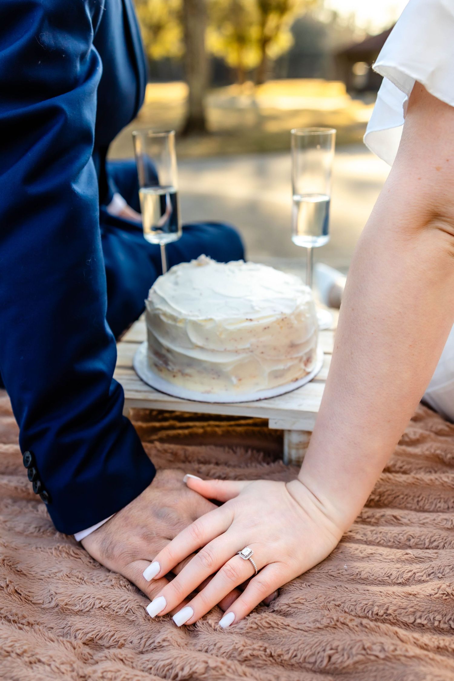 Close-up of hands touching near champagne glasses and a white cake on a textured blanket.