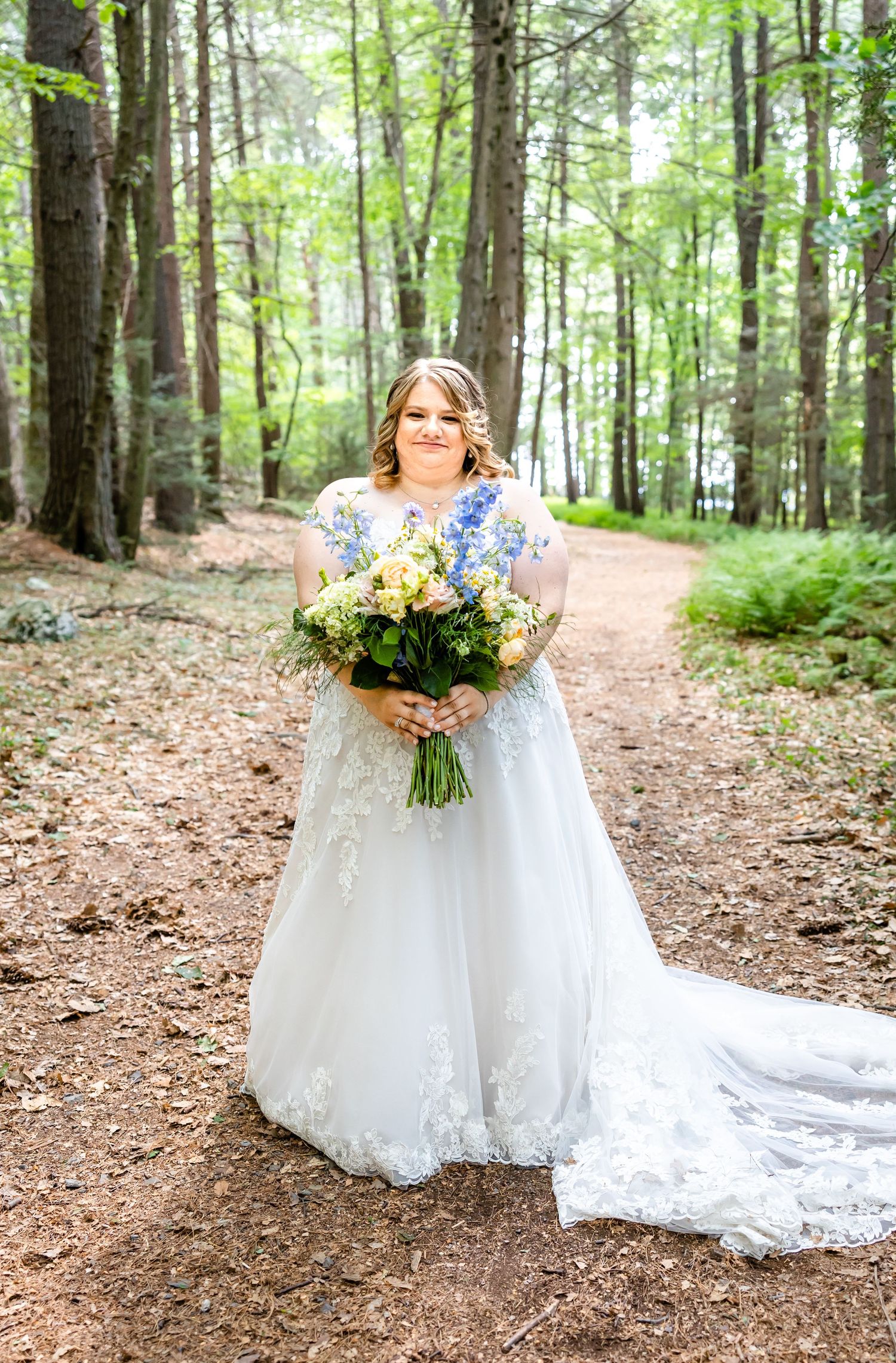 A bride in a lace wedding gown holds a colorful bouquet while standing on a forest path.