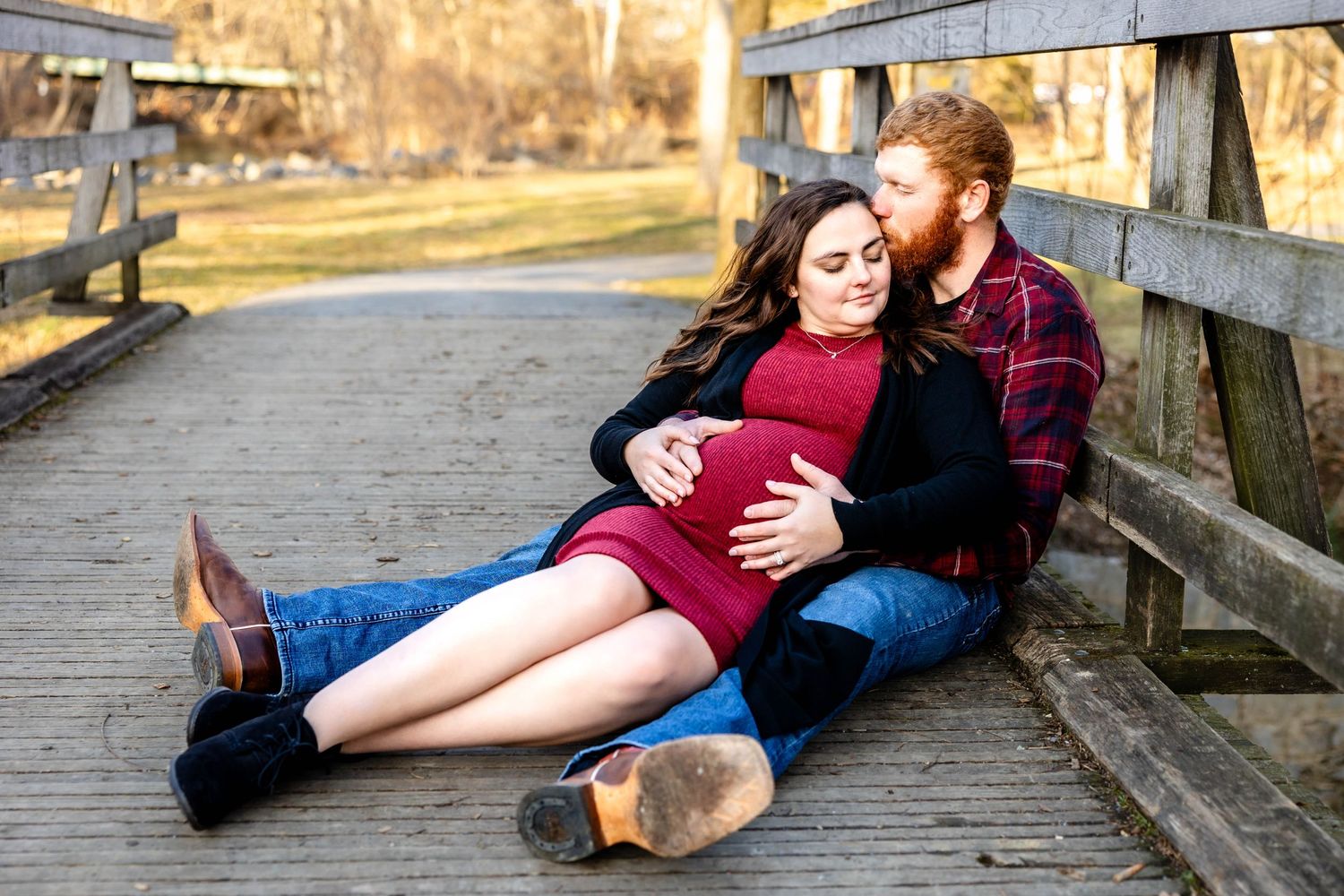 A couple relaxes on a wooden bridge during an outdoor maternity photo session.