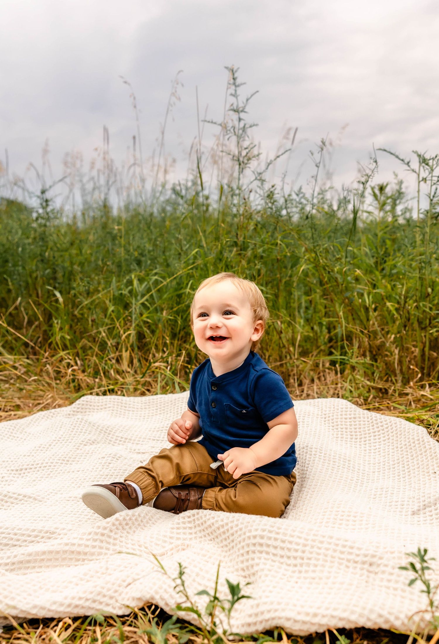 A toddler in navy blue shirt and khaki pants sits on white blanket surrounded by tall grass.