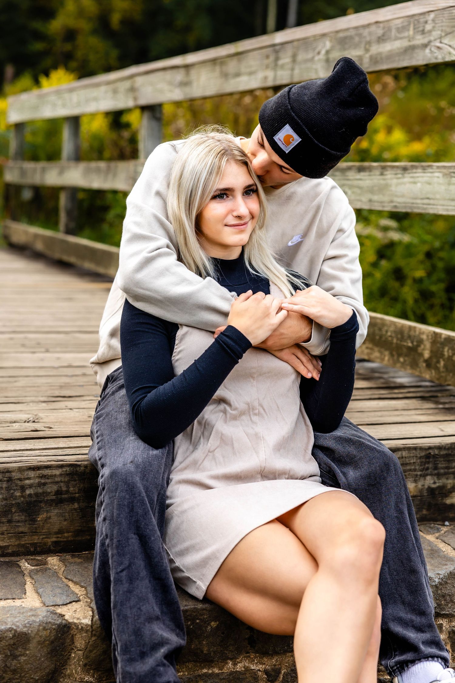 Couple sharing tender embrace on wooden bridge deck with fall colors in background.