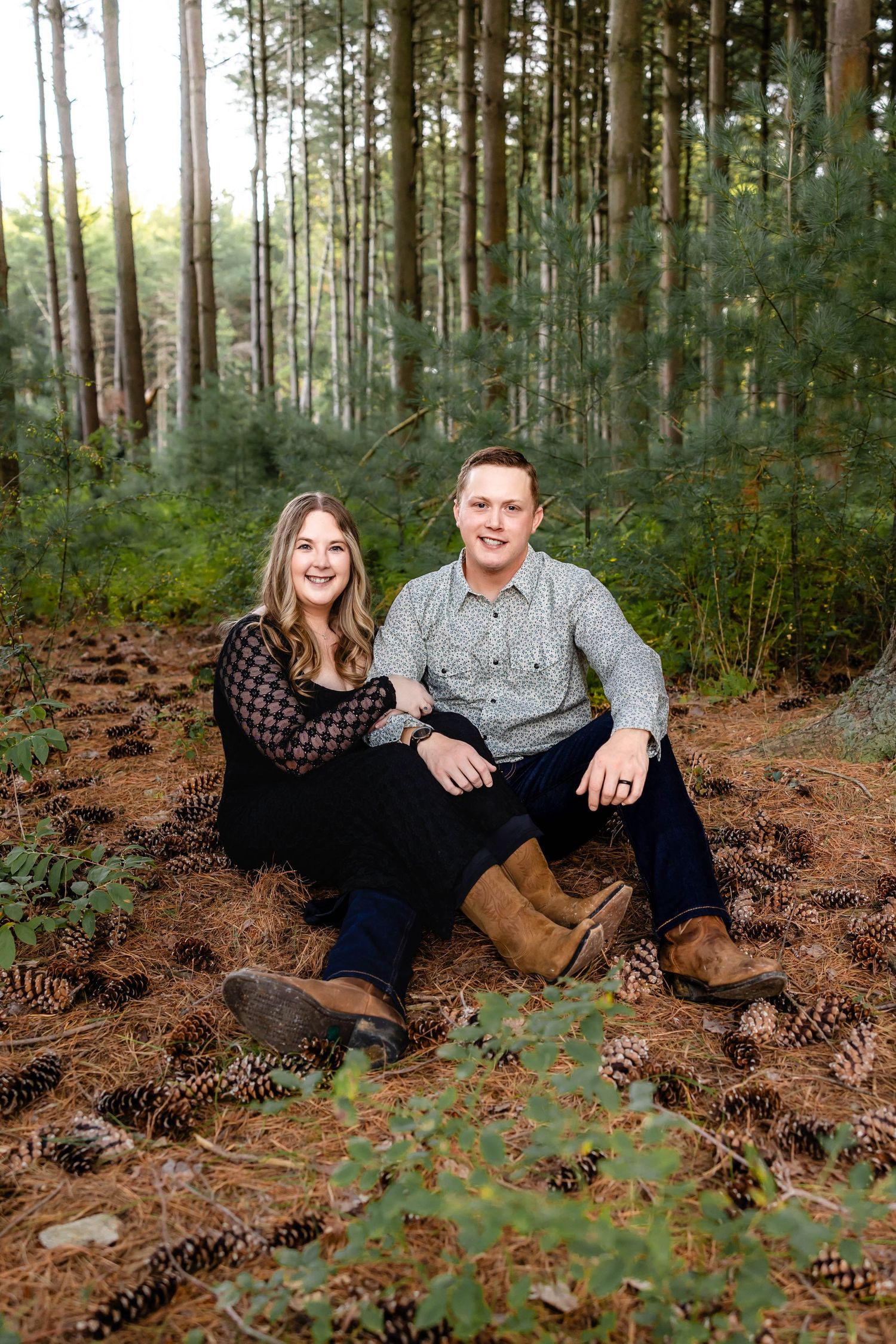 A couple sits together on pine needle-covered ground in a serene forest setting.