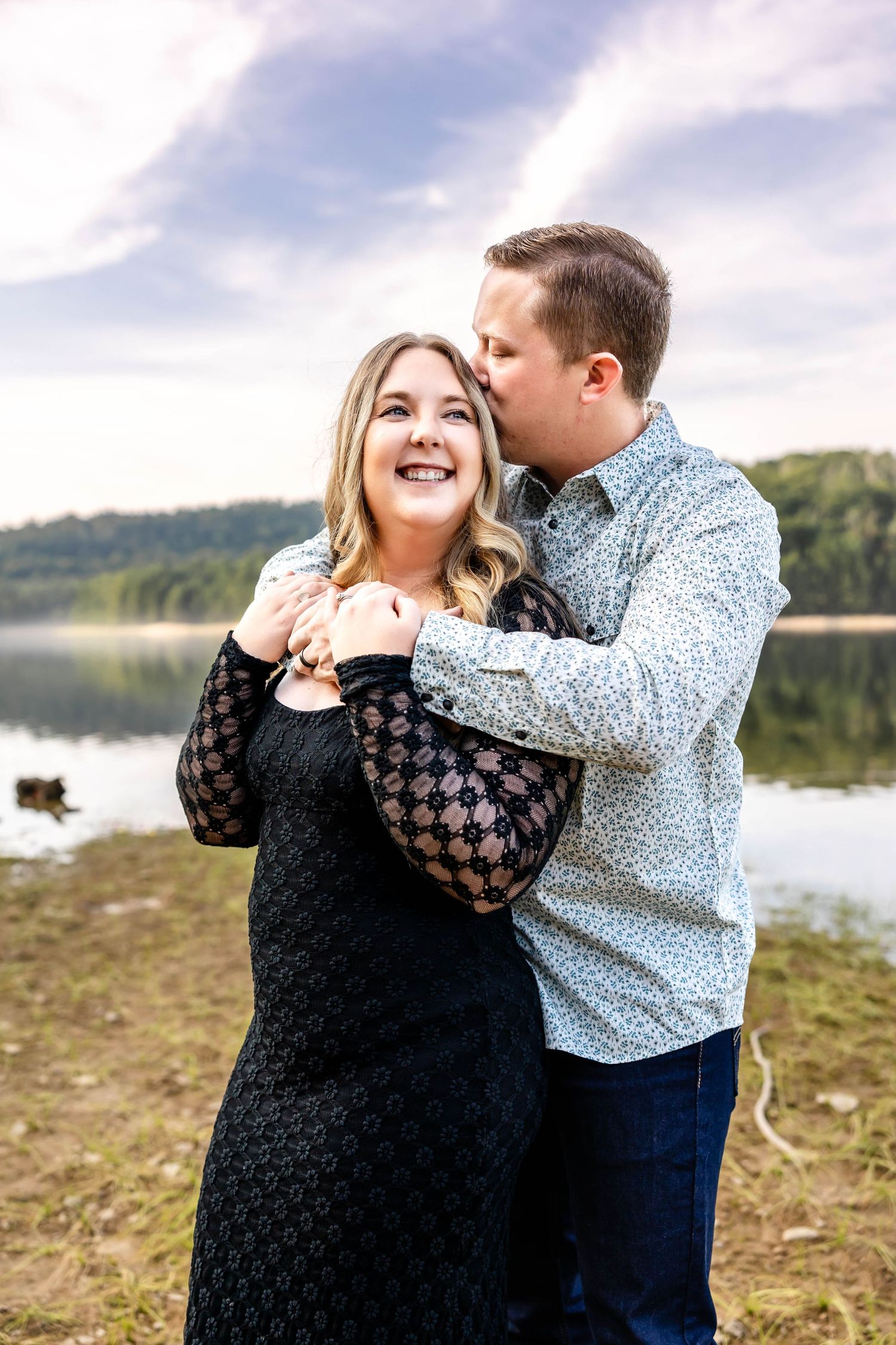 Romantic couple embraces by a serene lake with mountains in background during an engagement photoshoot.