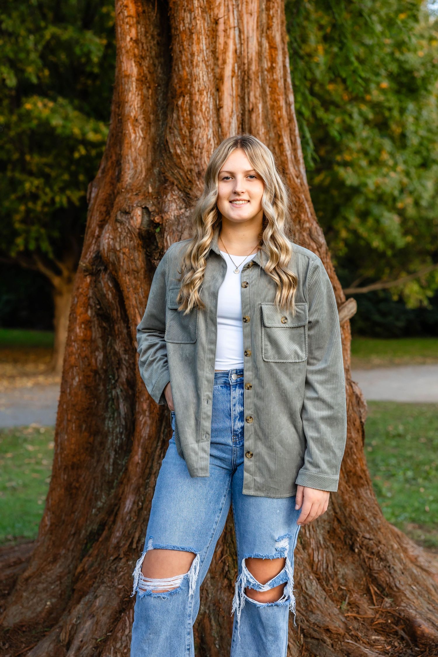 Young woman in gray jacket and ripped jeans poses in front of large redwood tree.