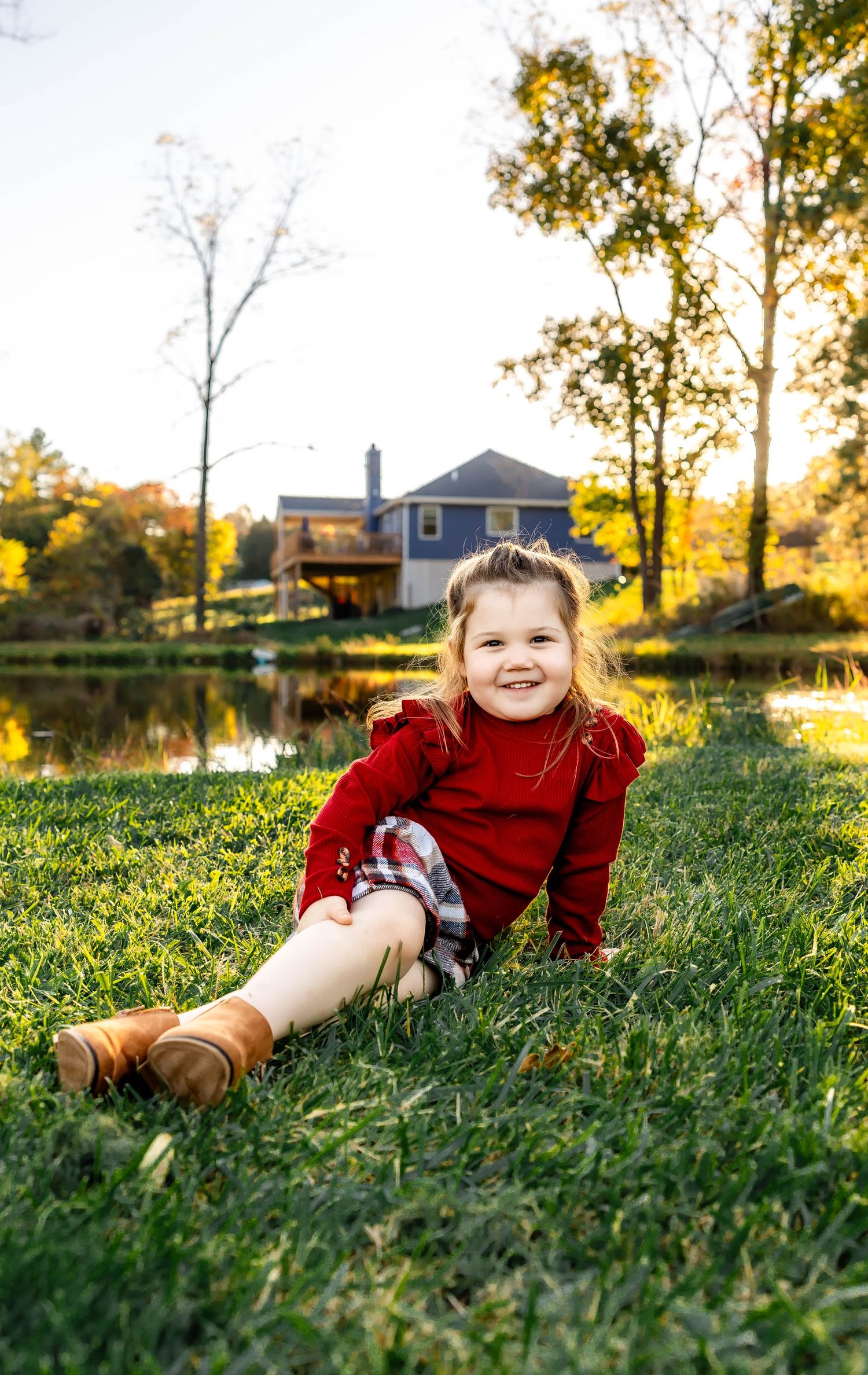 Child in red sweater and boots sits on grass with blue house and pond in autumn background.