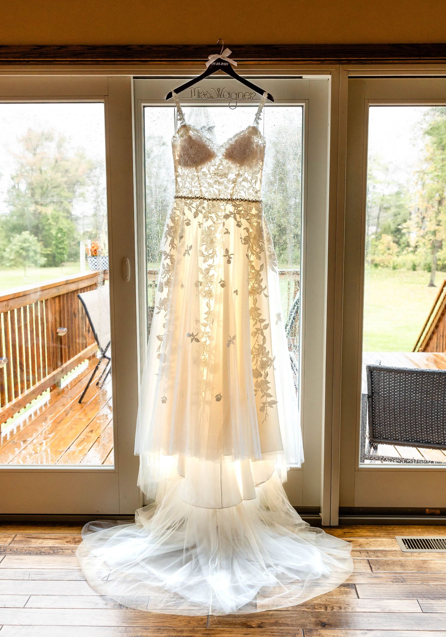 Elegant lace wedding dress hanging in window with deck view and natural lighting.