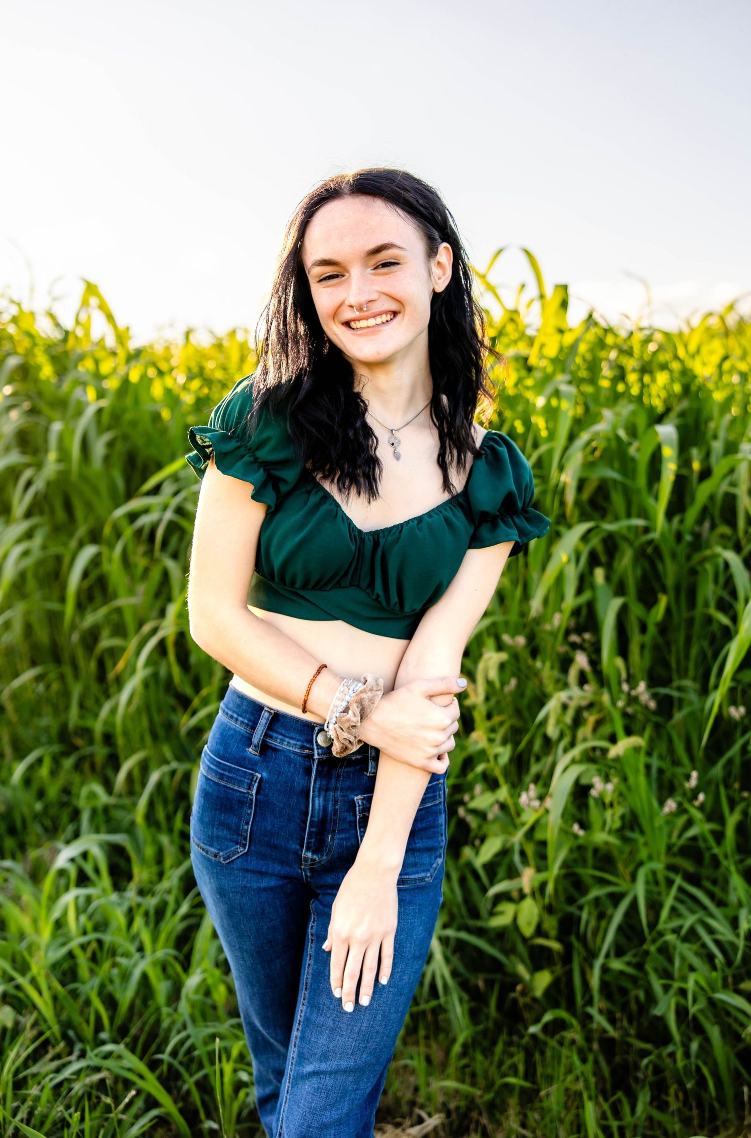 A person in a green crop top and jeans stands before a tall cornfield at sunset.
