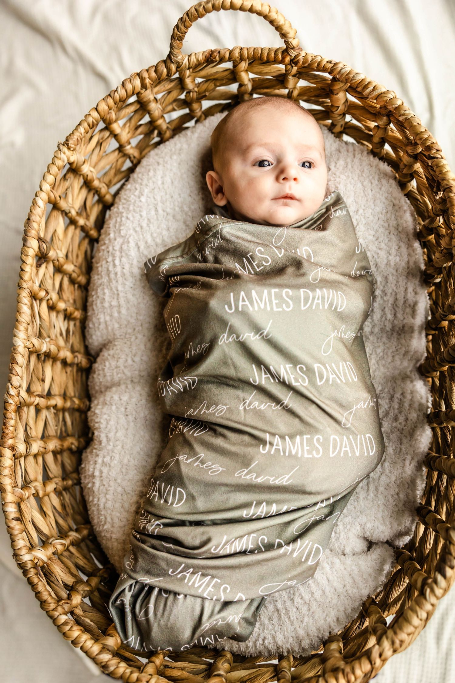 Baby swaddled in personalized gray blanket nestled in wicker basket on white bedding.