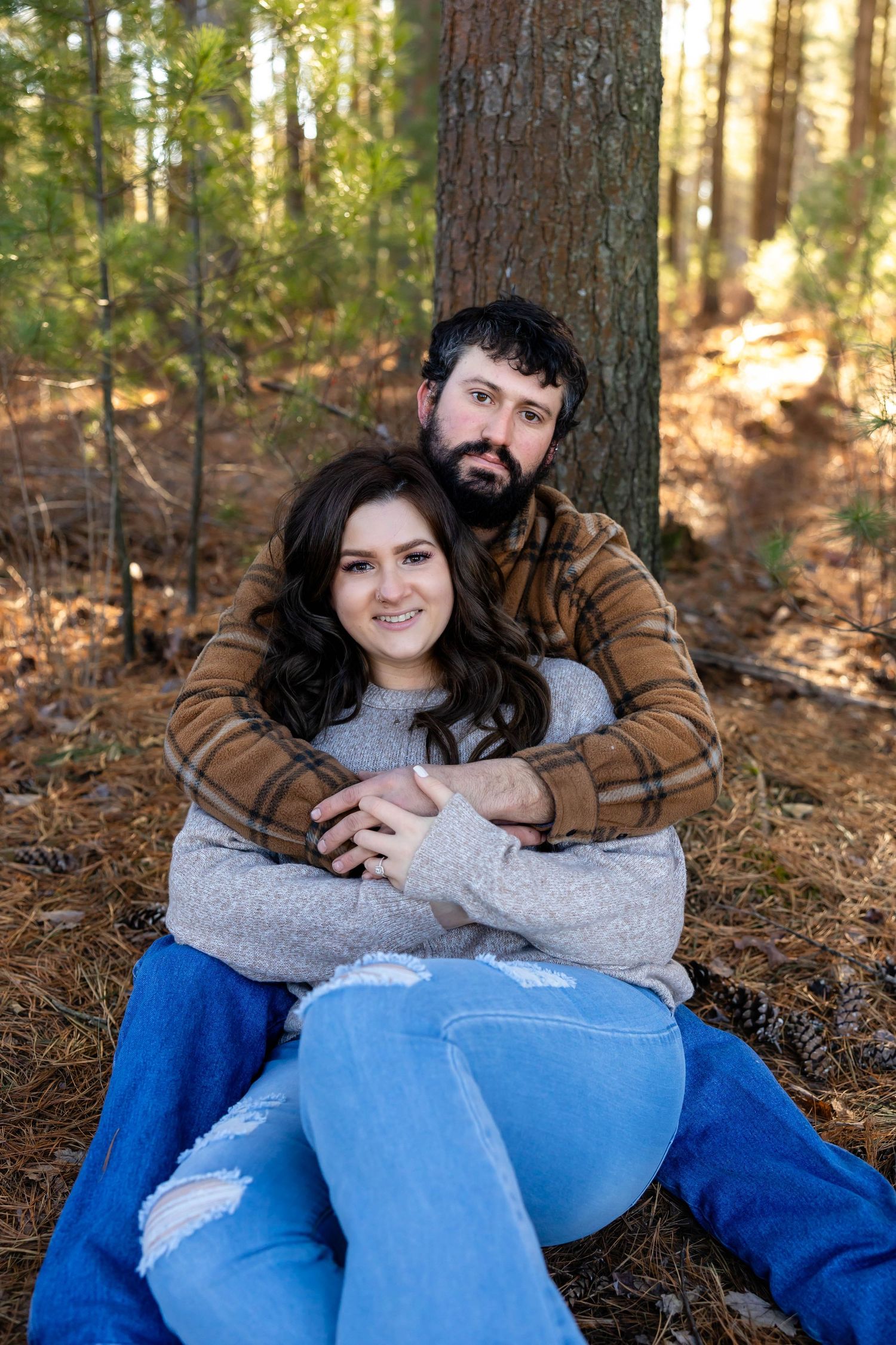 A couple sits together on fallen pine needles in a forest setting wearing casual autumn clothes.