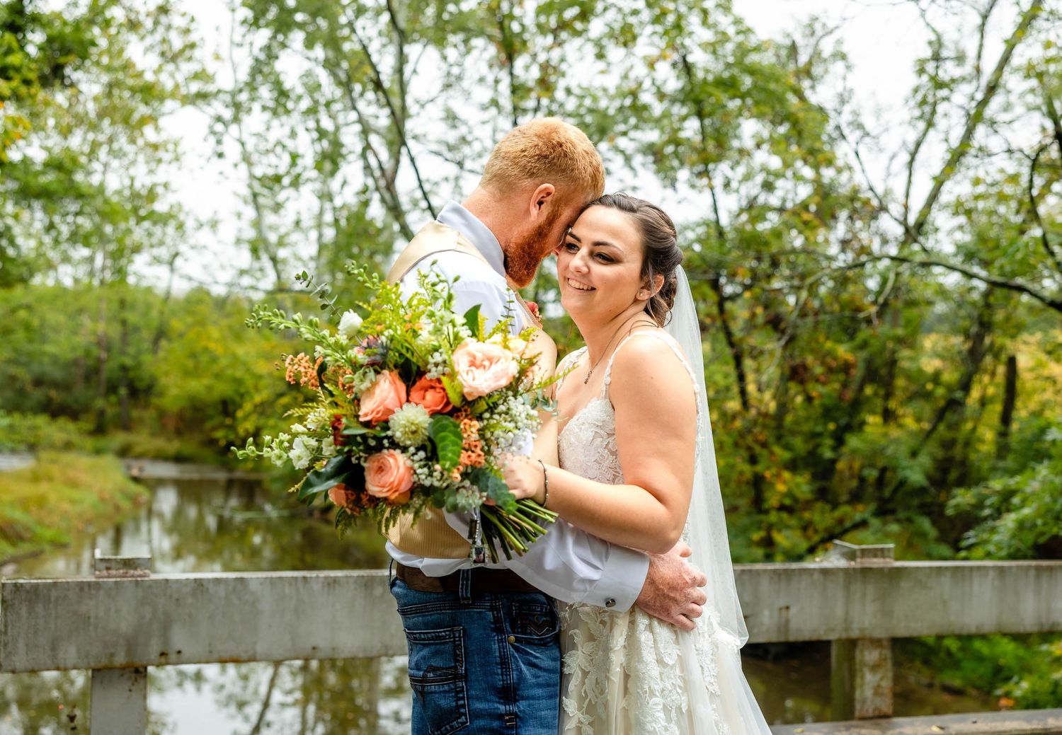 Happy wedding couple laughing together on rustic bridge with floral bouquet.