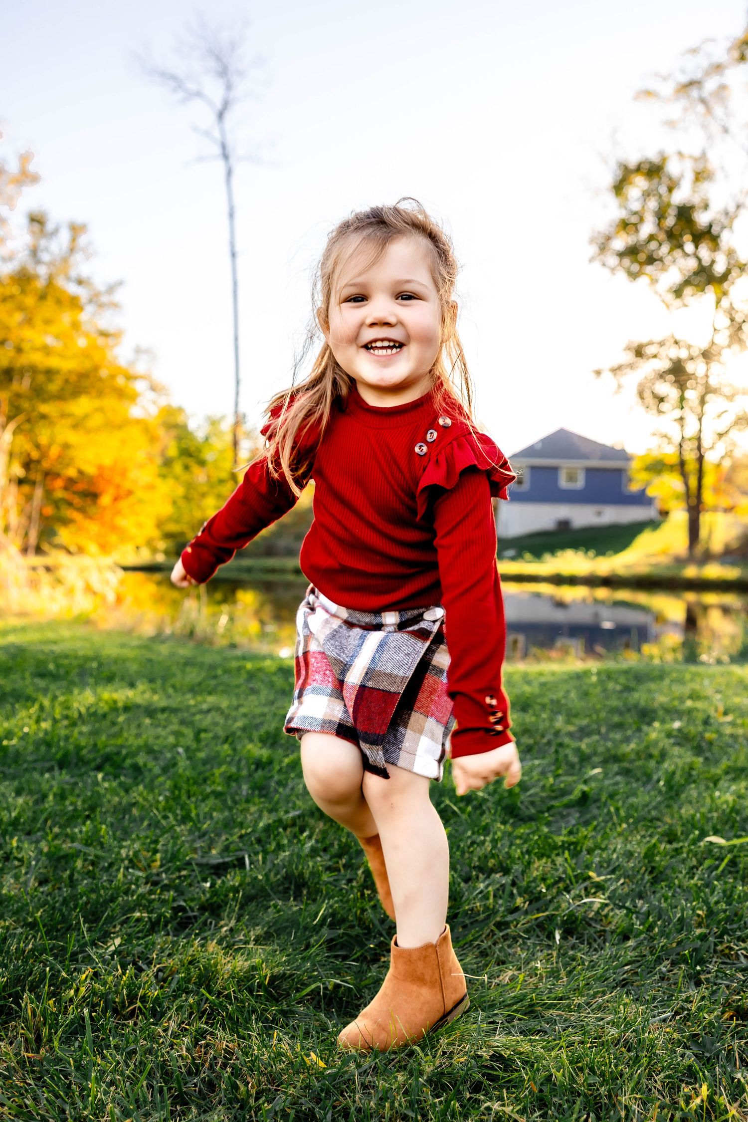 Child in red sweater and plaid skirt jumping joyfully in autumn garden setting.