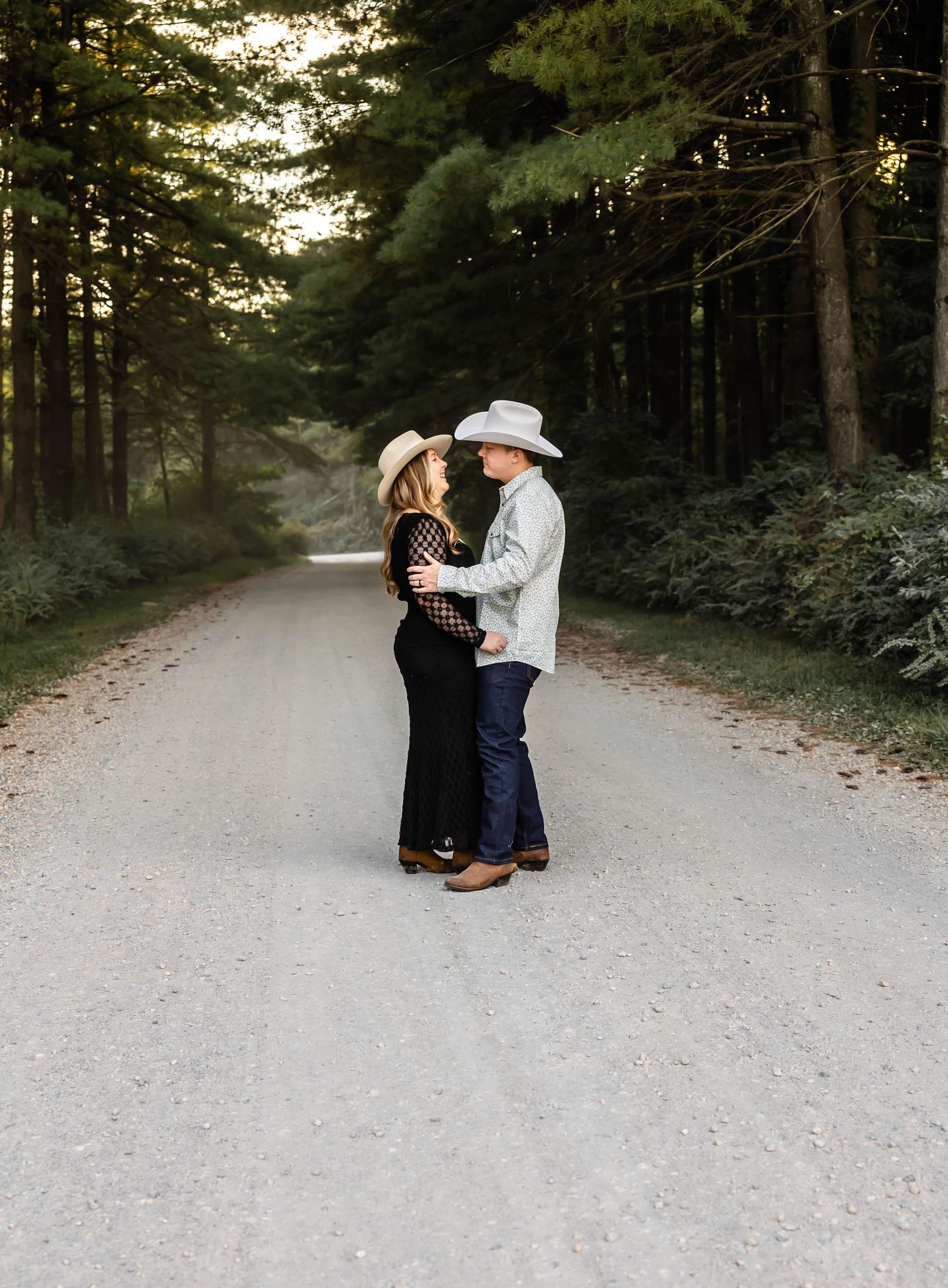 A couple in western attire embraces on a peaceful forest road.