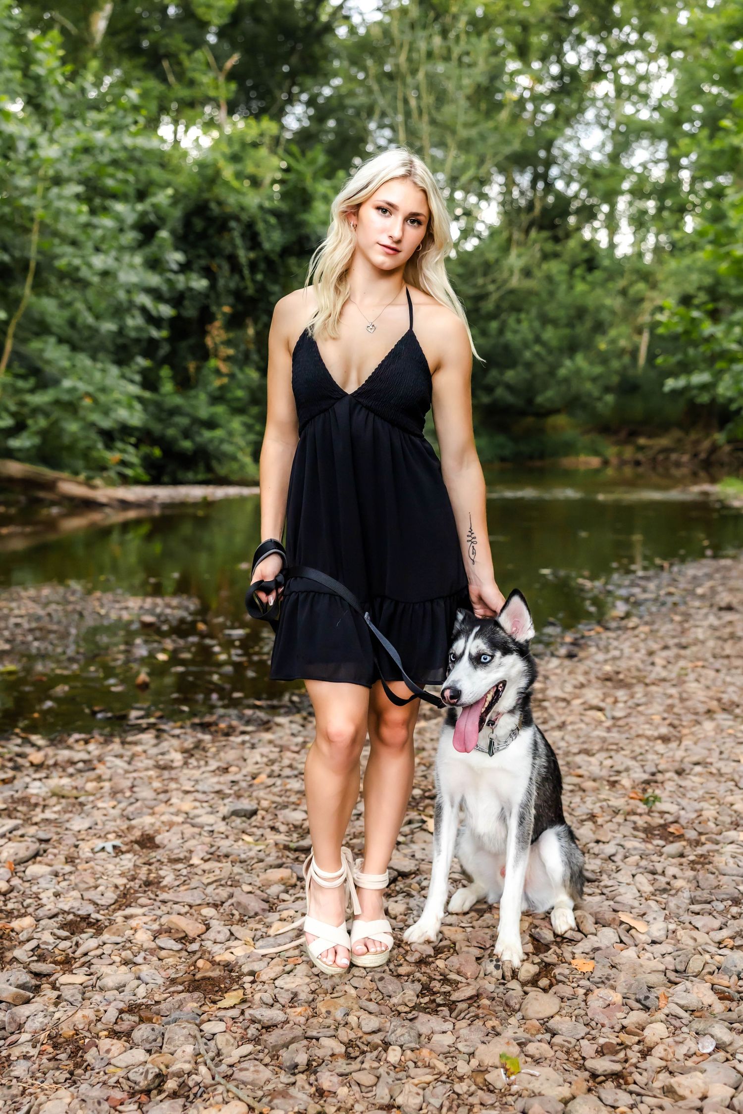 A black sundress-clad person stands with their happy Husky dog by a stream.