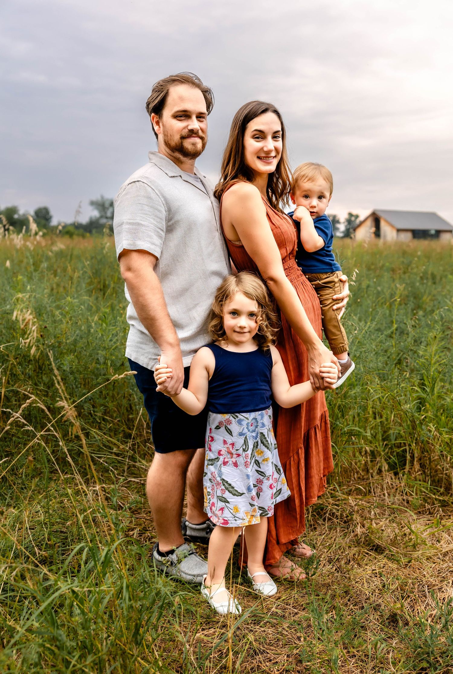 Family portrait in a rustic field setting with a farmhouse in the background.