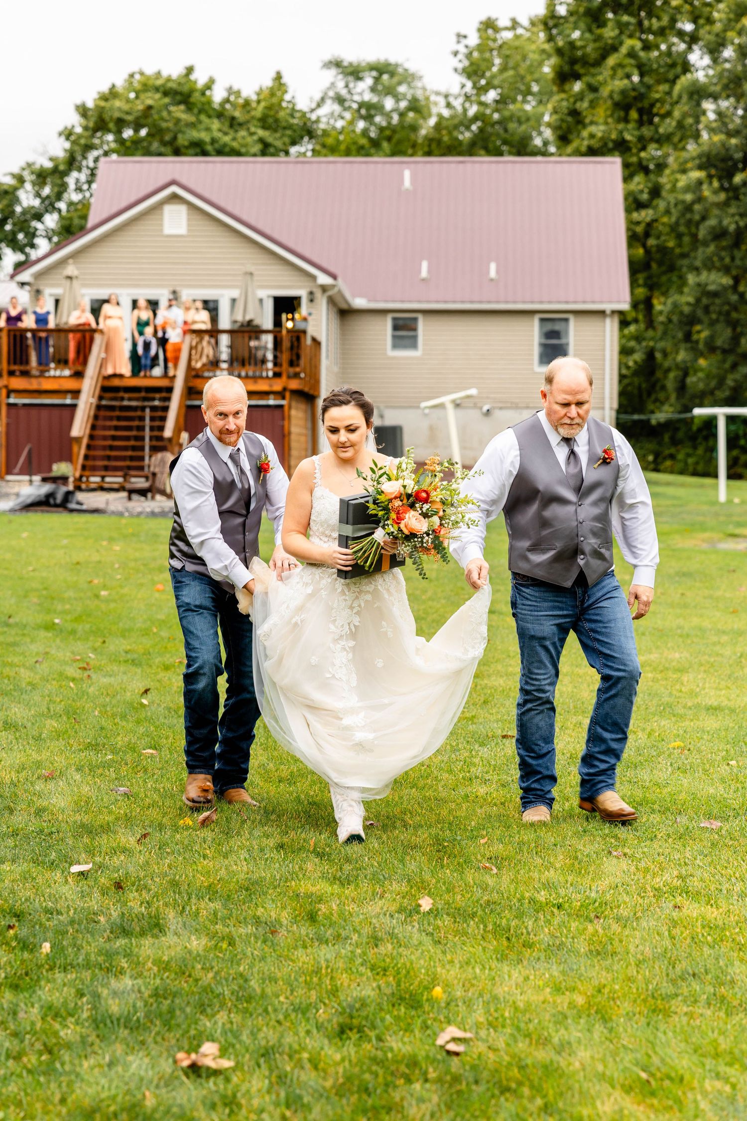 A couple in wedding attire runs across a lawn with groomsmen in gray vests in front of a house with a deck.